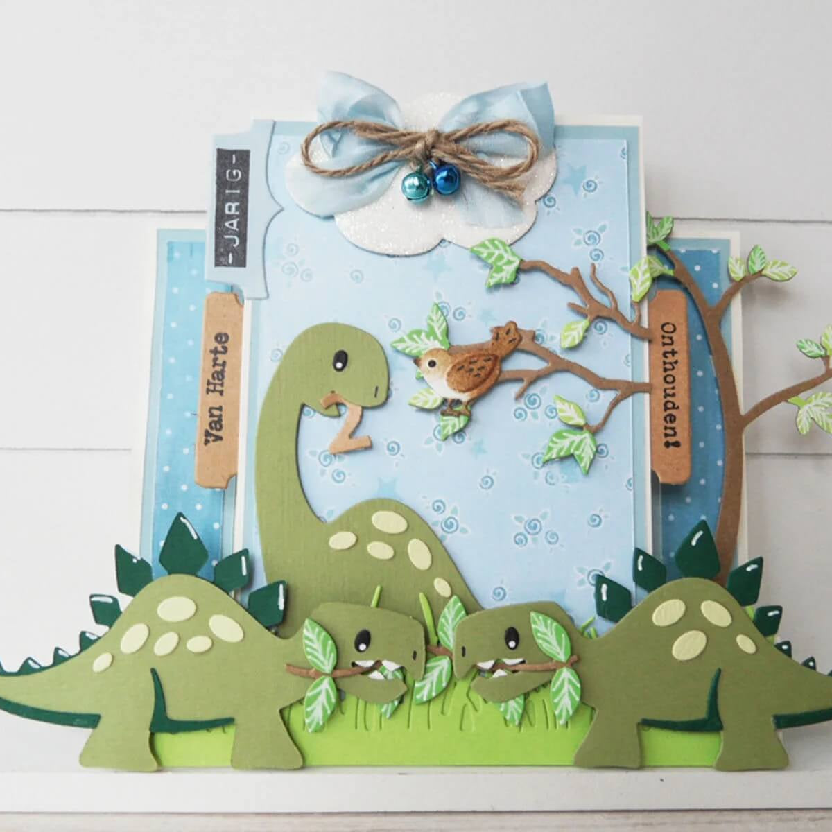 Handgefertigte Karte mit Marianne Design - Collectables Präge- und Stanzschablone Eline's Dinosaurier von Marianne Design, mit Cartoon-Dinosauriern, Blättern, Baum, Vogel, Wolke mit Schleife und Glöckchen; inklusive holländischen Grußanhängern. Ideal für Scrapbooking und Stanzmaschinen.