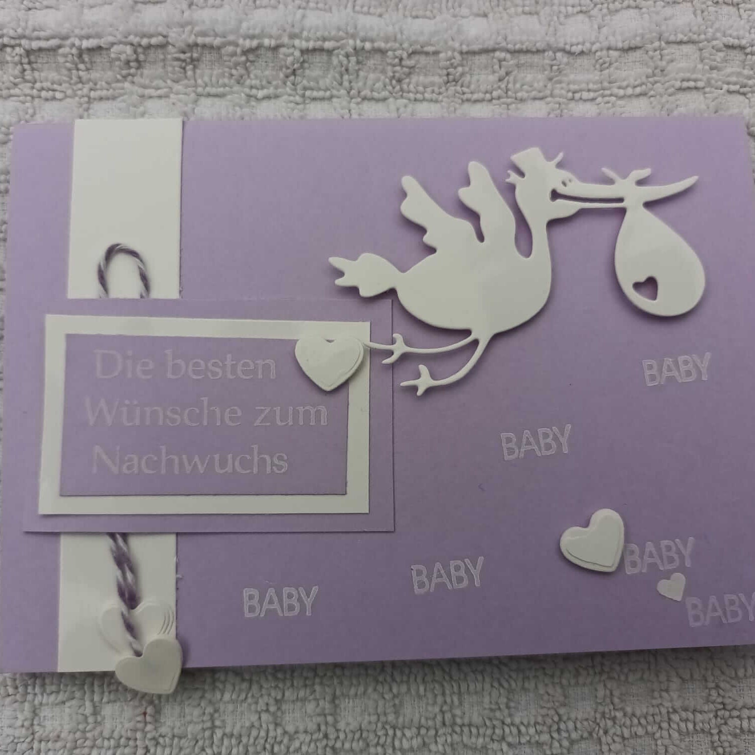 Eine lila Karte zeigt die Stanzschablone Storch mit Beutel im Schnabel von Stanzenshop.de, mit weißem Storch, Herzen und "Die besten Wünsche zum Nachwuchs" und "BABY" - ideal zum Basteln oder Scrapbooking auf strukturiertem Papier.