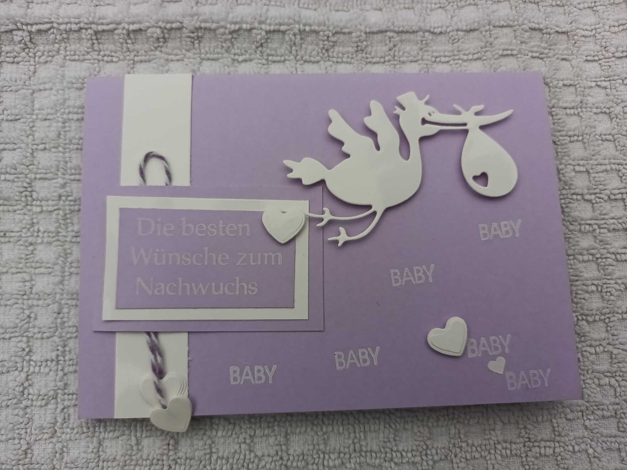 Eine lila Karte zeigt die Stanzschablone Storch mit Beutel im Schnabel von Stanzenshop.de, mit weißem Storch, Herzen und "Die besten Wünsche zum Nachwuchs" und "BABY" - ideal zum Basteln oder Scrapbooking auf strukturiertem Papier.