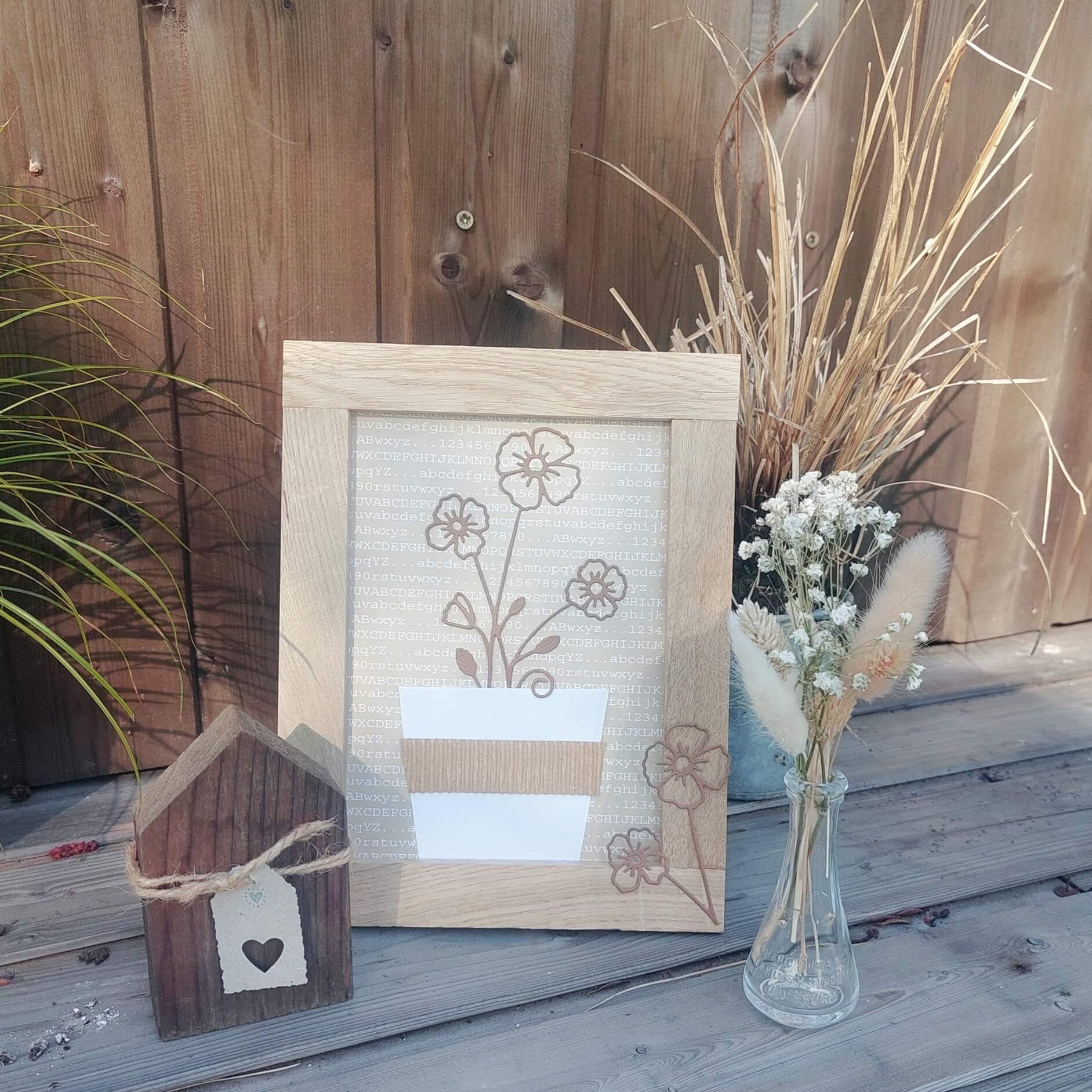 Ein gerahmter Blumendruck, ein kleines Holzhaus und eine Trockenblumenvase auf Holz bieten die perfekte Inspiration zum Basteln oder Ausstellen Ihrer Stanzschablone Blume mit drei Blüten von Stanzenshop.de-alles arrangiert vor einem Holzzaun.