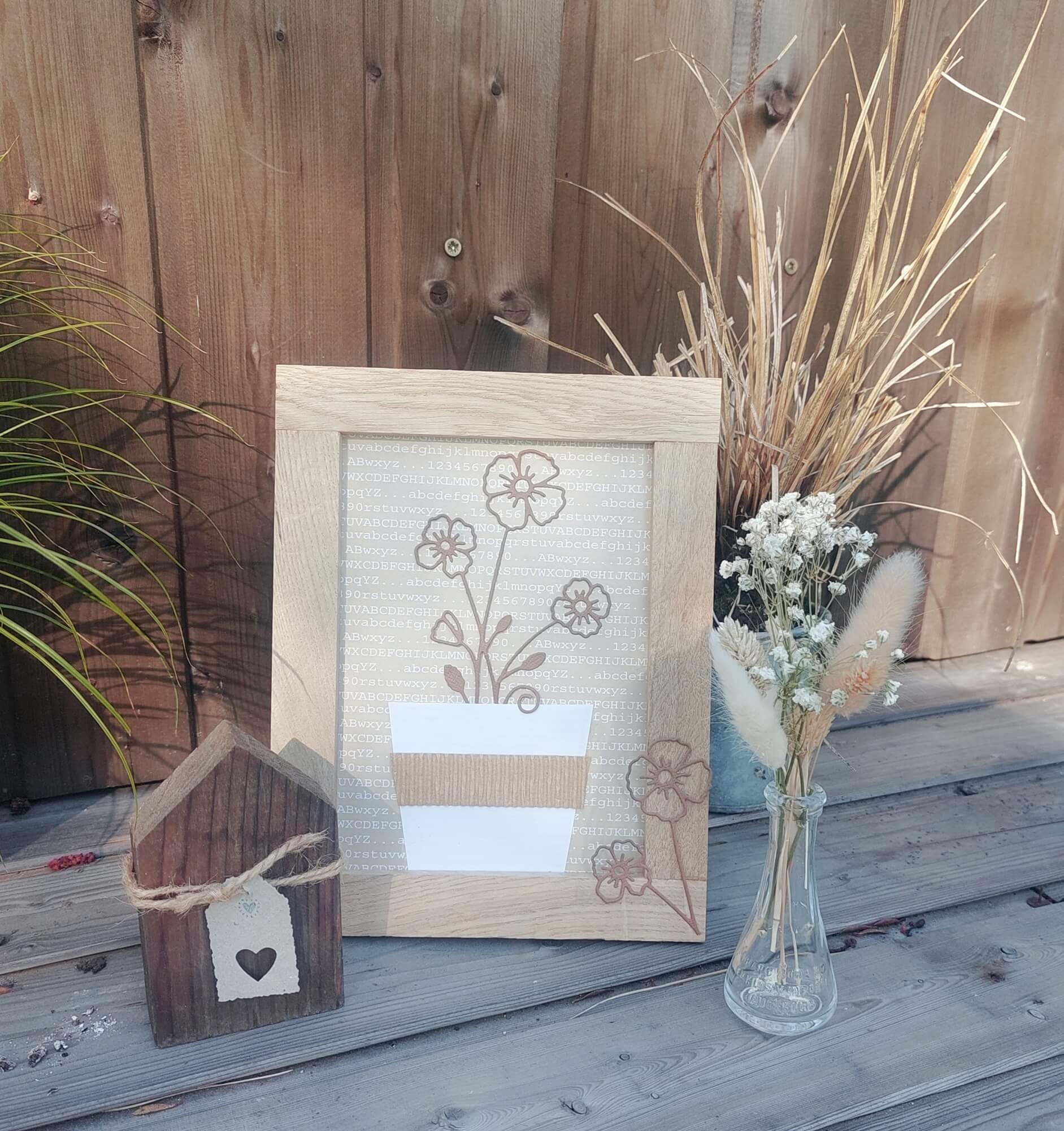 Ein gerahmter Blumendruck, ein kleines Holzhaus und eine Trockenblumenvase auf Holz bieten die perfekte Inspiration zum Basteln oder Ausstellen Ihrer Stanzschablone Blume mit drei Blüten von Stanzenshop.de-alles arrangiert vor einem Holzzaun.