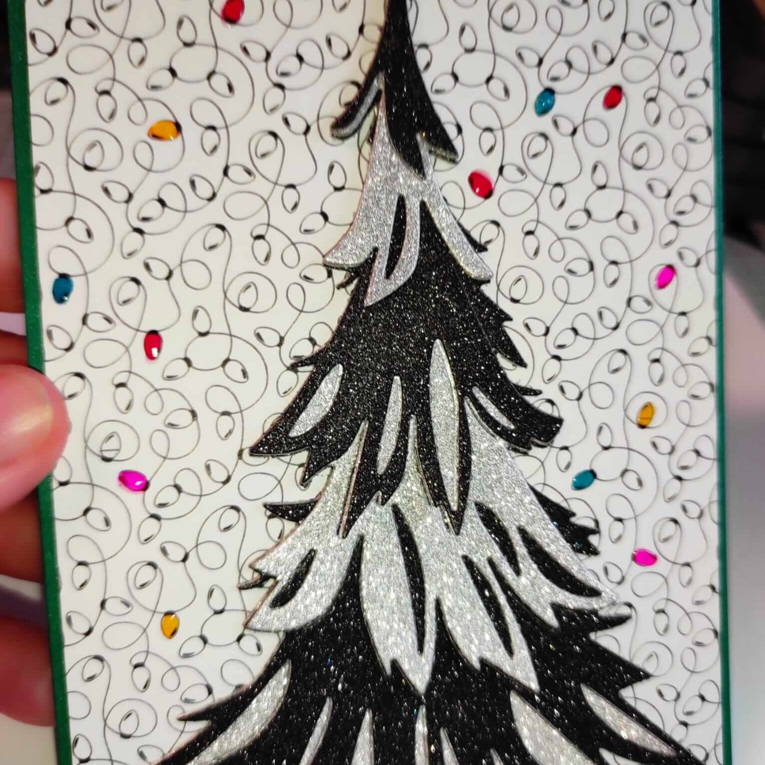 Eine Hand hält eine Karte mit einem schwarz-weißen Weihnachtsbaum, hergestellt mit den Sizzix 666332 - Thinlits Stanzschablonen Trim a Tree Colorize von Sizzix, auf einem gewirbelten Hintergrund mit bunten Punkten - perfekt für Weihnachten.