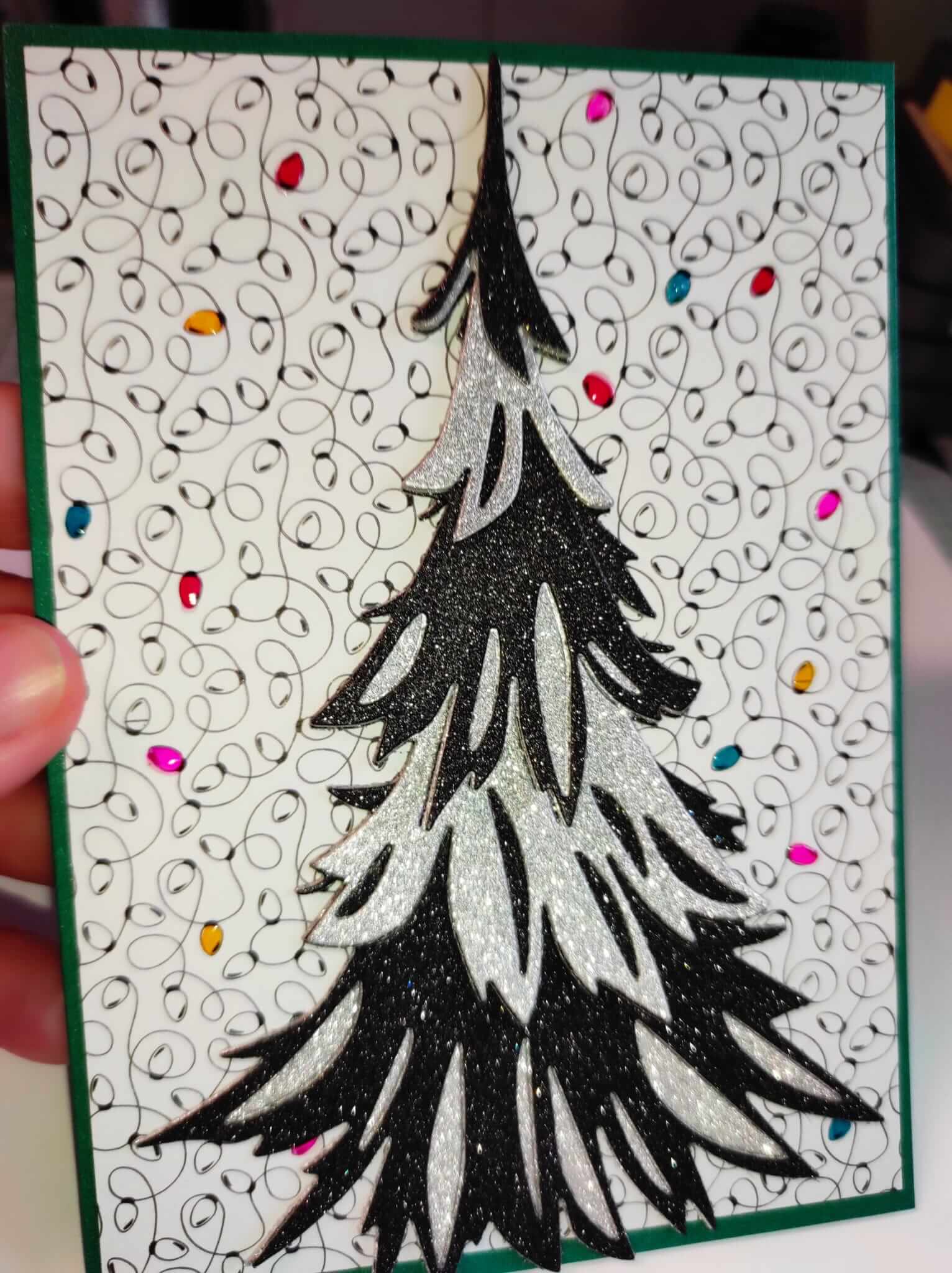 Eine Hand hält eine Karte mit einem schwarz-weißen Weihnachtsbaum, hergestellt mit den Sizzix 666332 - Thinlits Stanzschablonen Trim a Tree Colorize von Sizzix, auf einem gewirbelten Hintergrund mit bunten Punkten - perfekt für Weihnachten.