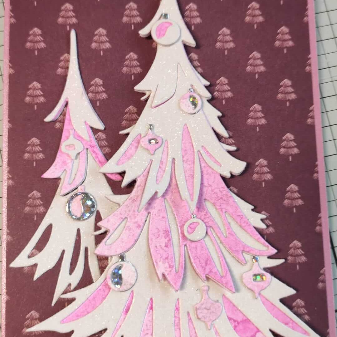 Handgemachte Karte mit geschichteten weißen und rosafarbenen Weihnachtsbäumen und Ornamenten, hergestellt mit den Sizzix 666332 Thinlits Stanzschablonen Trim a Tree Colorize von Sizzix, auf einem lila Hintergrund mit kleinen Baummustern - ideal für Weihnachten.