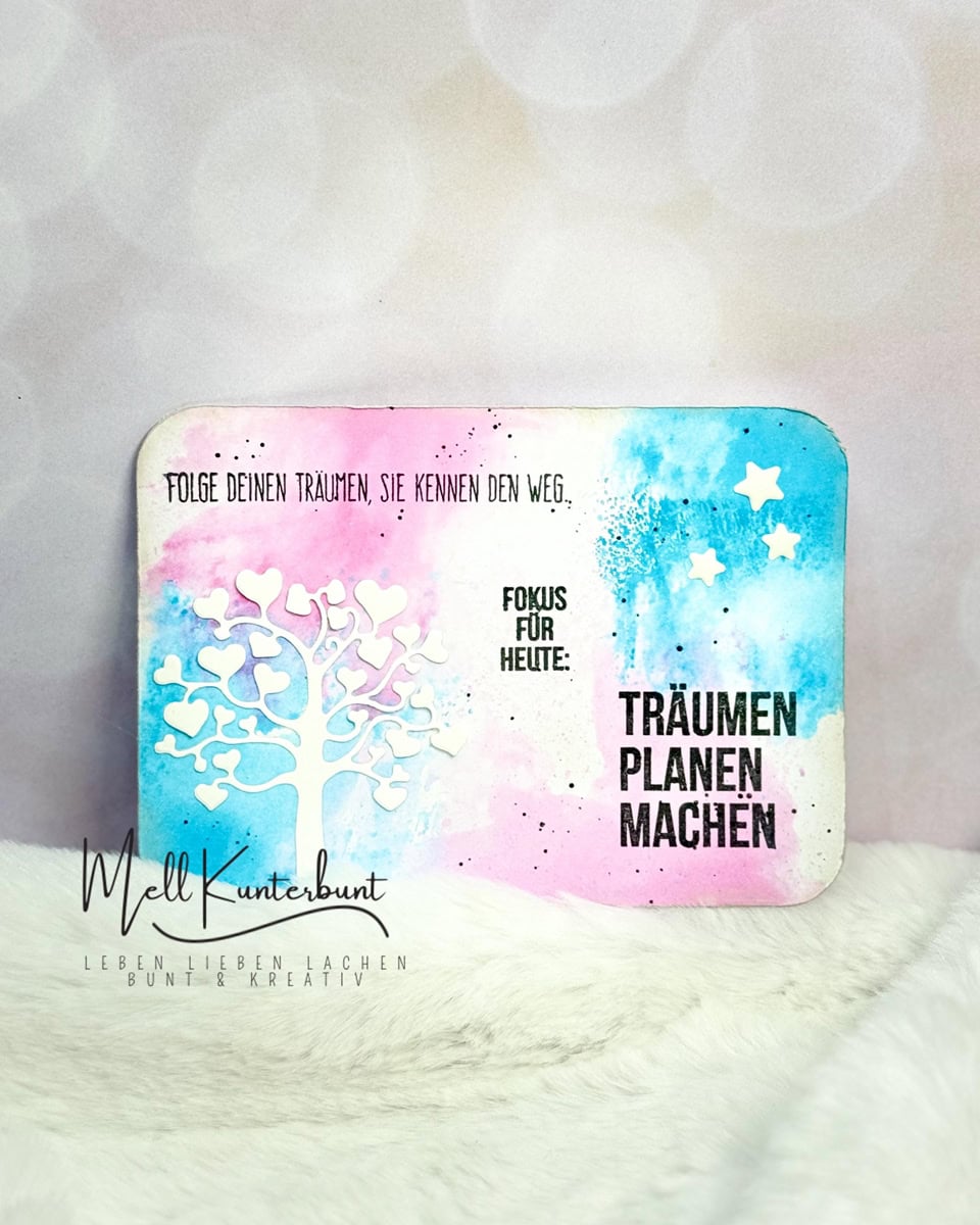 Eine lebendige Karte mit einer weißen Baumsilhouette aus der Stanzschablone: Baum mit Herzen von Stanzenshop.de, mit Sternausschnitten und deutschem Motivationstext auf blau-rosa Aquarellhintergrund - ideal für Scrapbooking oder Bastelzubehör.