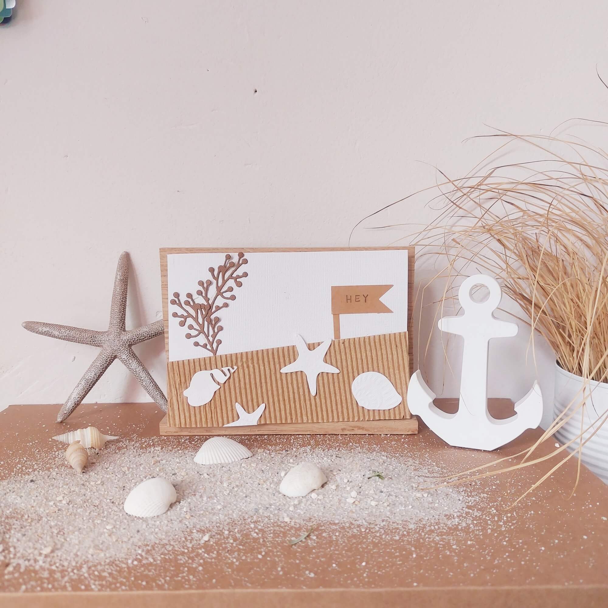 Ein dekoratives Arrangement mit Muscheln, einem Seestern, einem weißen Anker, getrocknetem Gras und einer Grußkarte mit maritimen Stanzschablonen Perlenzweige von Stanzenshop.de auf sandy decor.