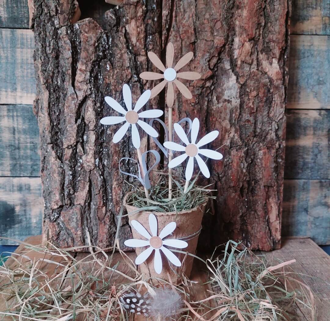 Die Stanzschablone: Blumenblüte von Stanzenshop.de zeigt zarte weiße und beigefarbene Gänseblümchenmotive, ideal zum Basteln oder für Scrapbooking-Projekte. Ausgestellt mit Heu auf Holzrinde für einen rustikalen Touch.