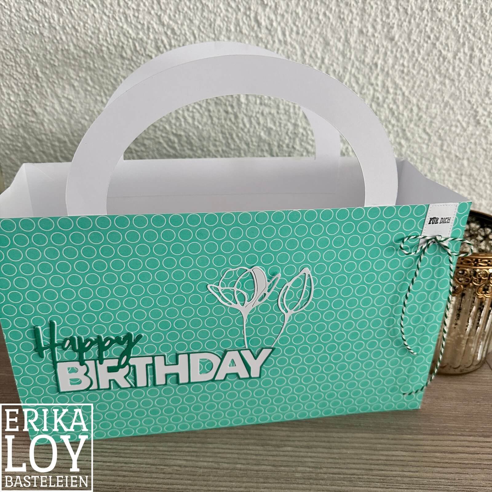 Eine tealfarbene, gemusterte Geschenktüte mit "Happy Birthday" und weißen Blumenmustern steht auf einem Tisch neben einem kleinen Glasbehälter, der perfekt zu den handgefertigten Karten der Stanzenshop.de Stanzschablone Blume mit zwei Blüten passt.