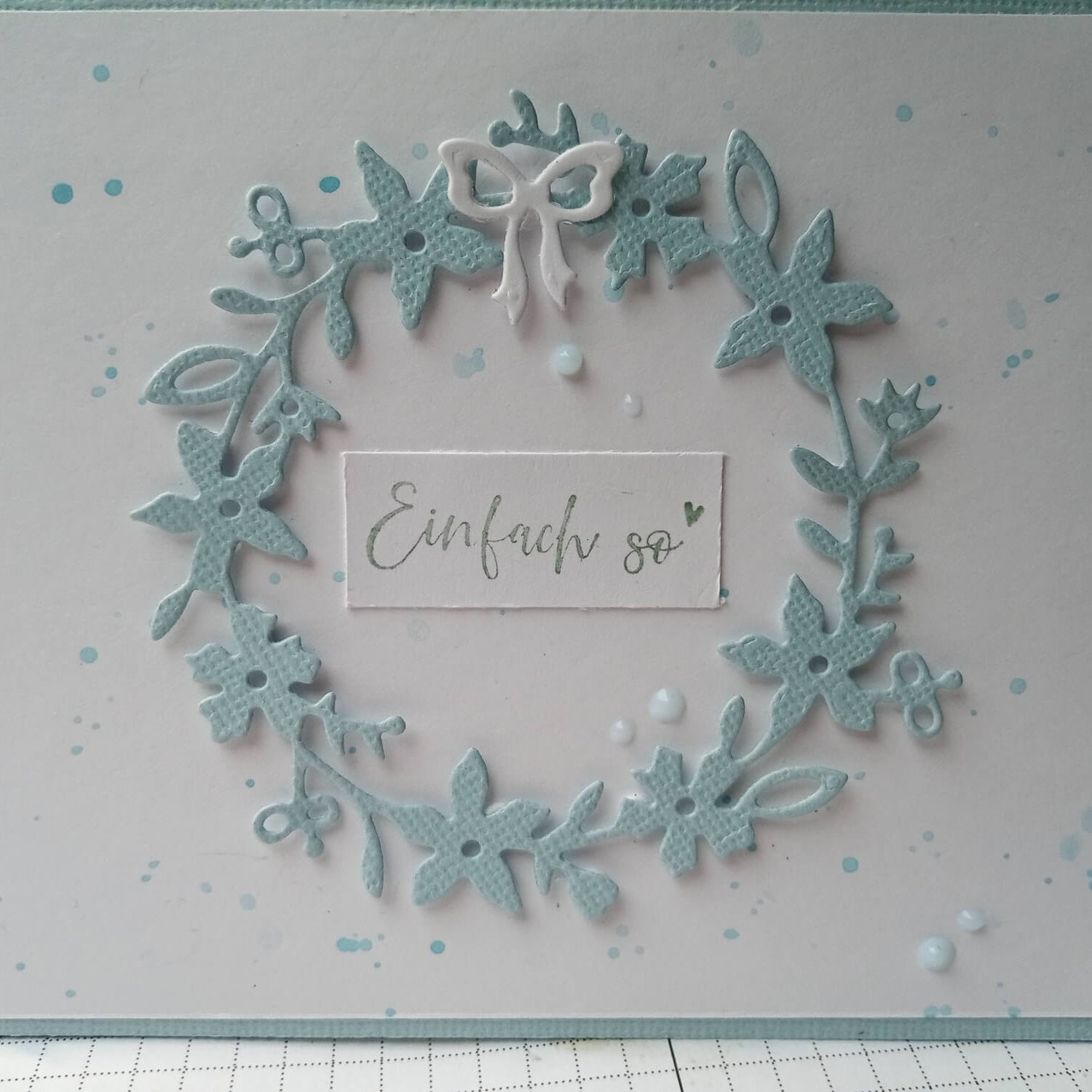 Eine handgemachte Karte mit der Stanzschablone Blumenkranz von Stanzenshop.de, verziert mit einem blauen Kranz, einer kleinen weißen Schleife und "Einfach so" auf weißem Hintergrund mit blauen Farbspritzern.