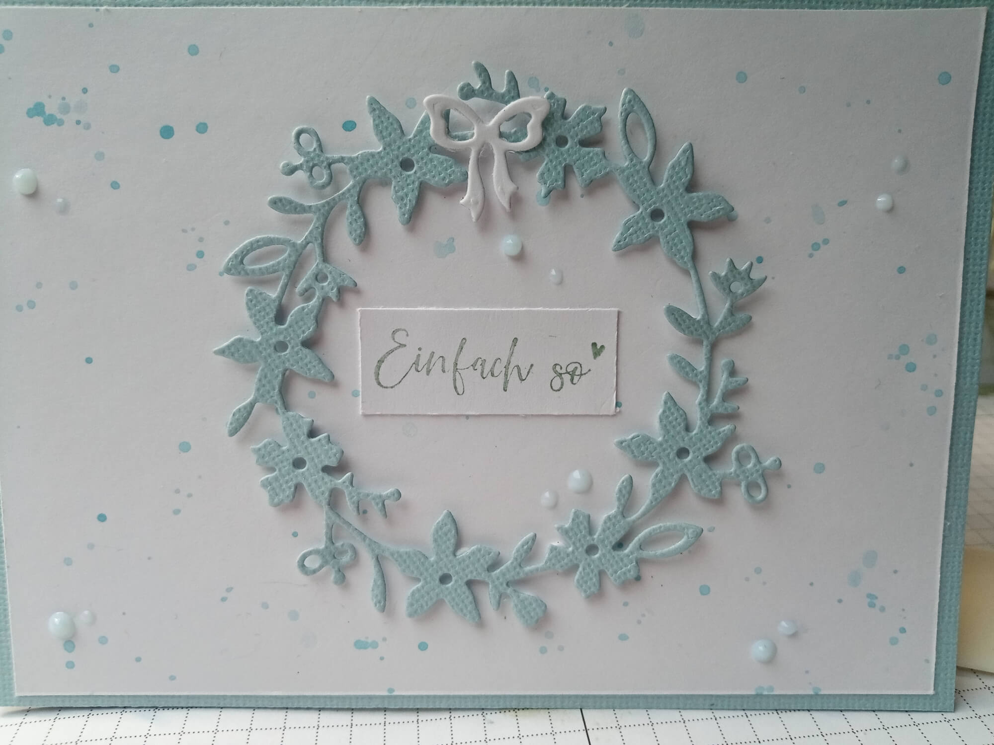 Eine handgemachte Karte mit der Stanzschablone Blumenkranz von Stanzenshop.de, verziert mit einem blauen Kranz, einer kleinen weißen Schleife und "Einfach so" auf weißem Hintergrund mit blauen Farbspritzern.