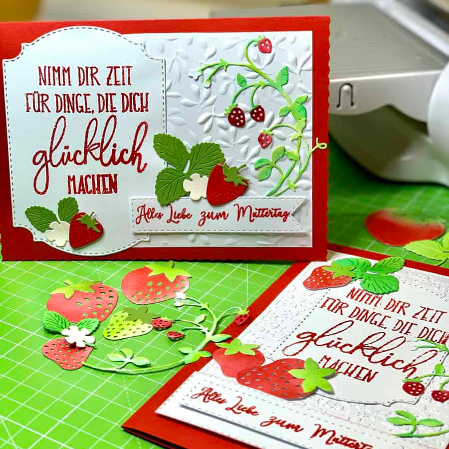Zwei handgefertigte Grußkarten mit Erdbeeren und Blättern, hergestellt mit der "Stanzschablone Erdbeeren" von Stanzenshop.de, mit deutschem Text auf einer grünen Schneidematte mit Bastelwerkzeug im Hintergrund.