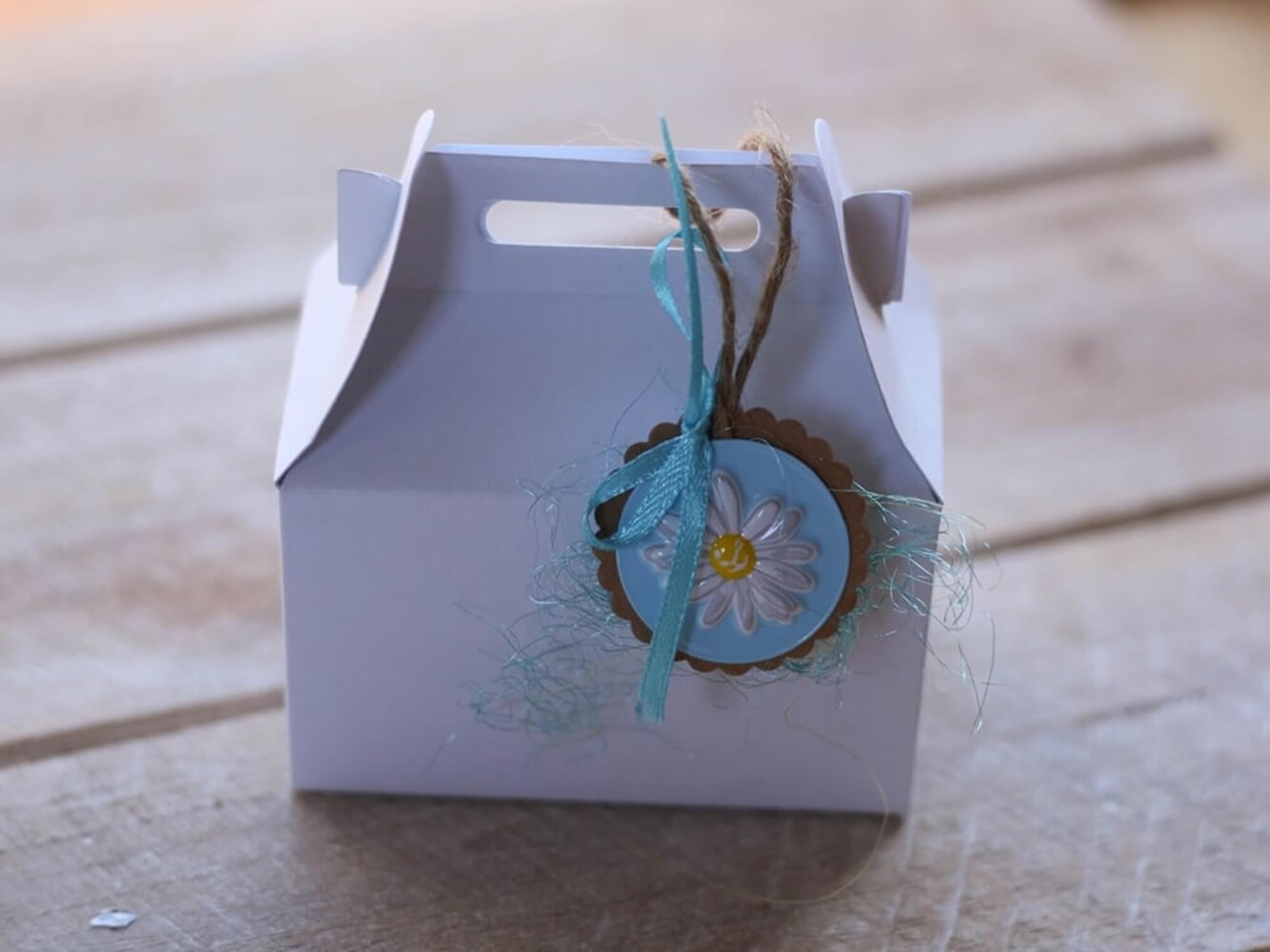 Eine kleine weiße Schachtel aus der Stanzschablone Große Schachtel von Stanzenshop.de mit Henkel, blauem Band, Schnur und blau-weißem Blumenanhänger steht auf einer Holzfläche.
