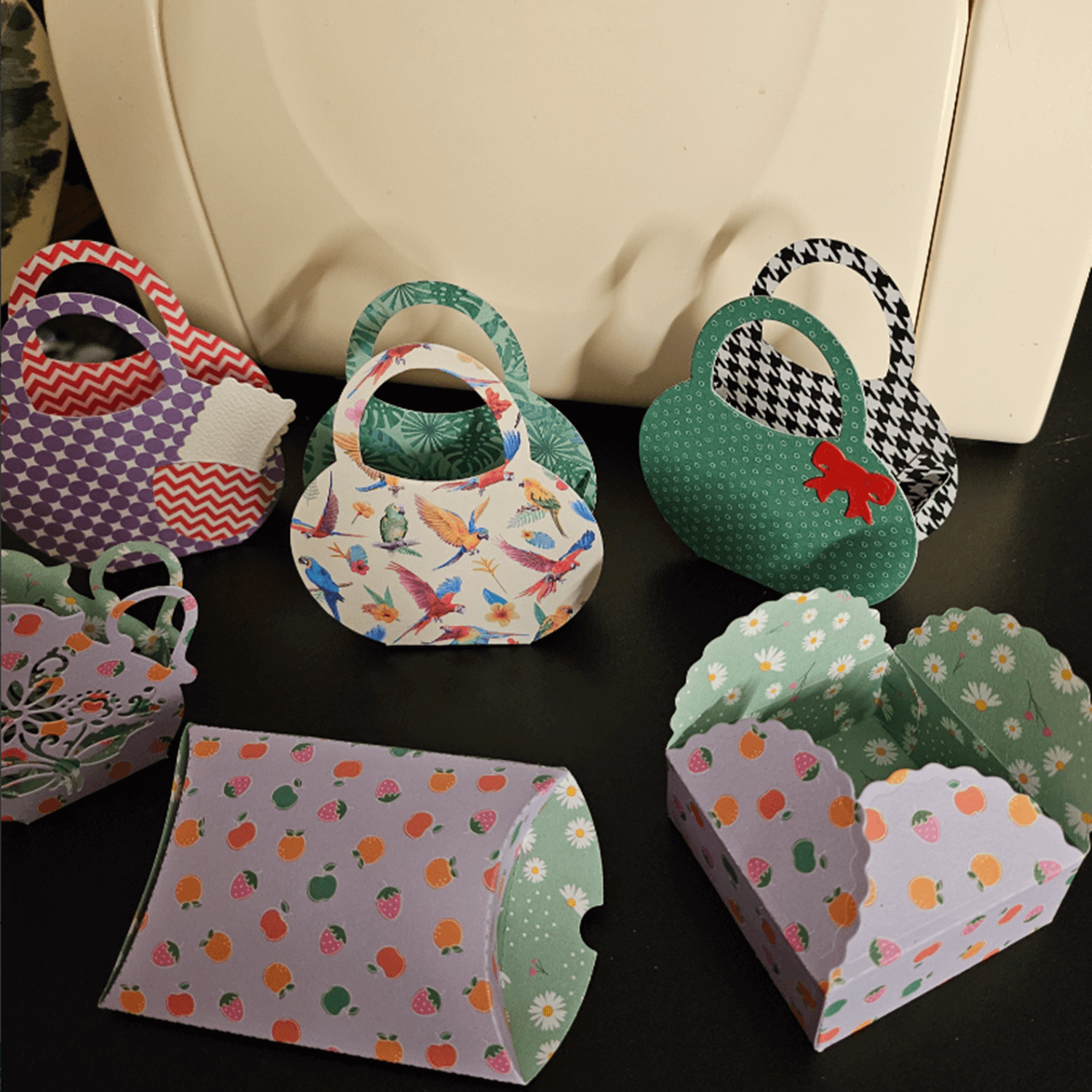 Stanzschablone Geschenkkästchen von Stanzenshop.de: Bunte, kleine Papier-Geschenkkästchen mit verschiedenen Mustern und Formen - ideal zum Basteln oder Scrapbooking - auf dunkler Fläche vor weißem Behälter ausgestellt.