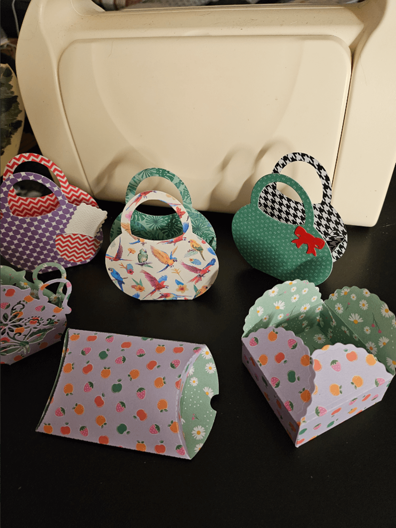 Stanzschablone Geschenkkästchen von Stanzenshop.de: Bunte, kleine Papier-Geschenkkästchen mit verschiedenen Mustern und Formen - ideal zum Basteln oder Scrapbooking - auf dunkler Fläche vor weißem Behälter ausgestellt.