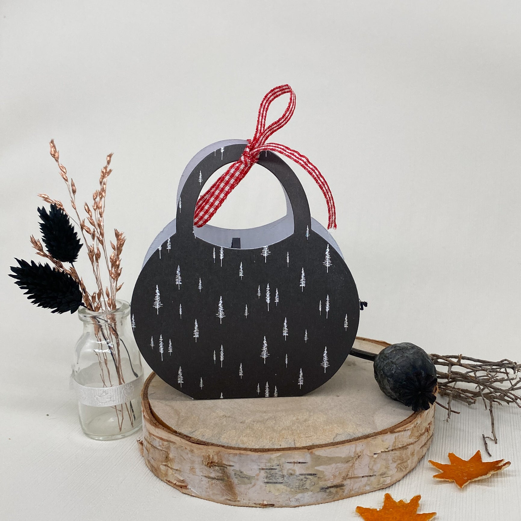 Eine schwarze, runde Papiertüte mit weißen Baummotiven und rotem Band aus der Stanzschablone Bastelset Handtasche von Stanzenshop.de liegt auf einer Holzplatte zwischen Herbstblättern und Trockenblumen - ideal zum Basteln oder Scrapbooking.
