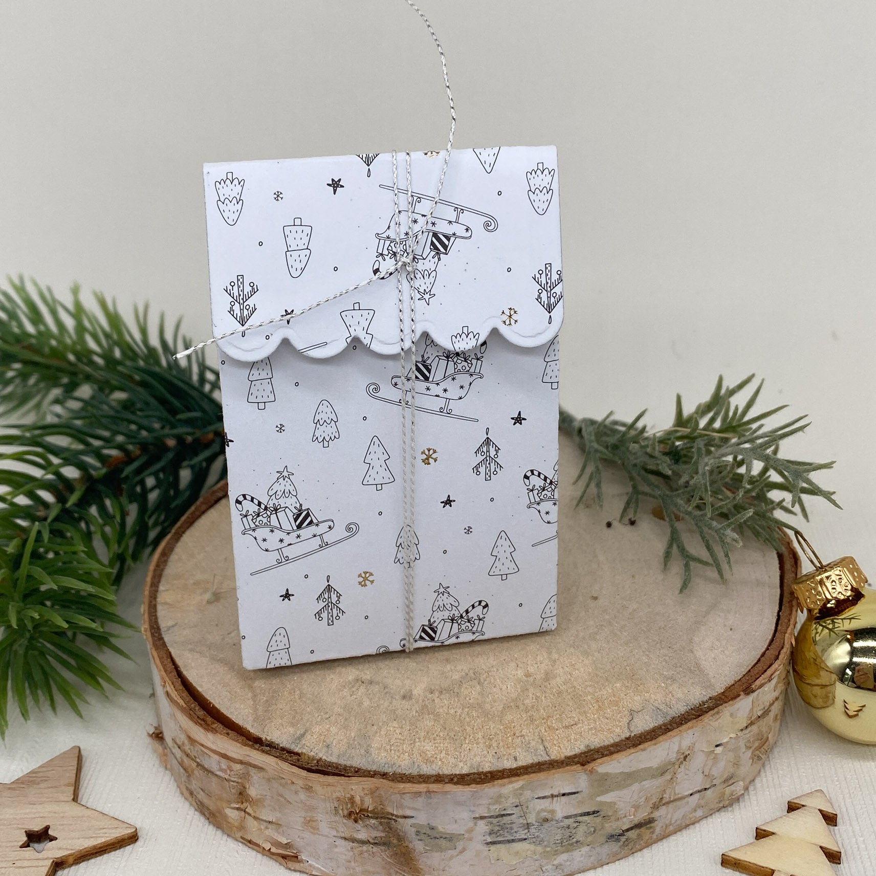 Ein weißer Geschenkumschlag mit winterlichen Motiven und Kordelverschluss ist auf Holz mit Tannenzweigen und Sternen abgebildet - die perfekte Inspiration für Gastgeschenke oder Bastelboxen mit der "Stanzschablone Schachtel mit Verzierungen" von Stanzenshop.de.