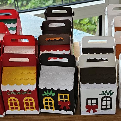 Ein Set bunter, hausförmiger Geschenkboxen mit dem Stanzschablone Bastelset für eine Schachtel mit winterlichen Motiven von Stanzenshop.de, mit einzigartigen Dach- und Türmotiven, arrangiert auf einem Tisch an einem Fenster.