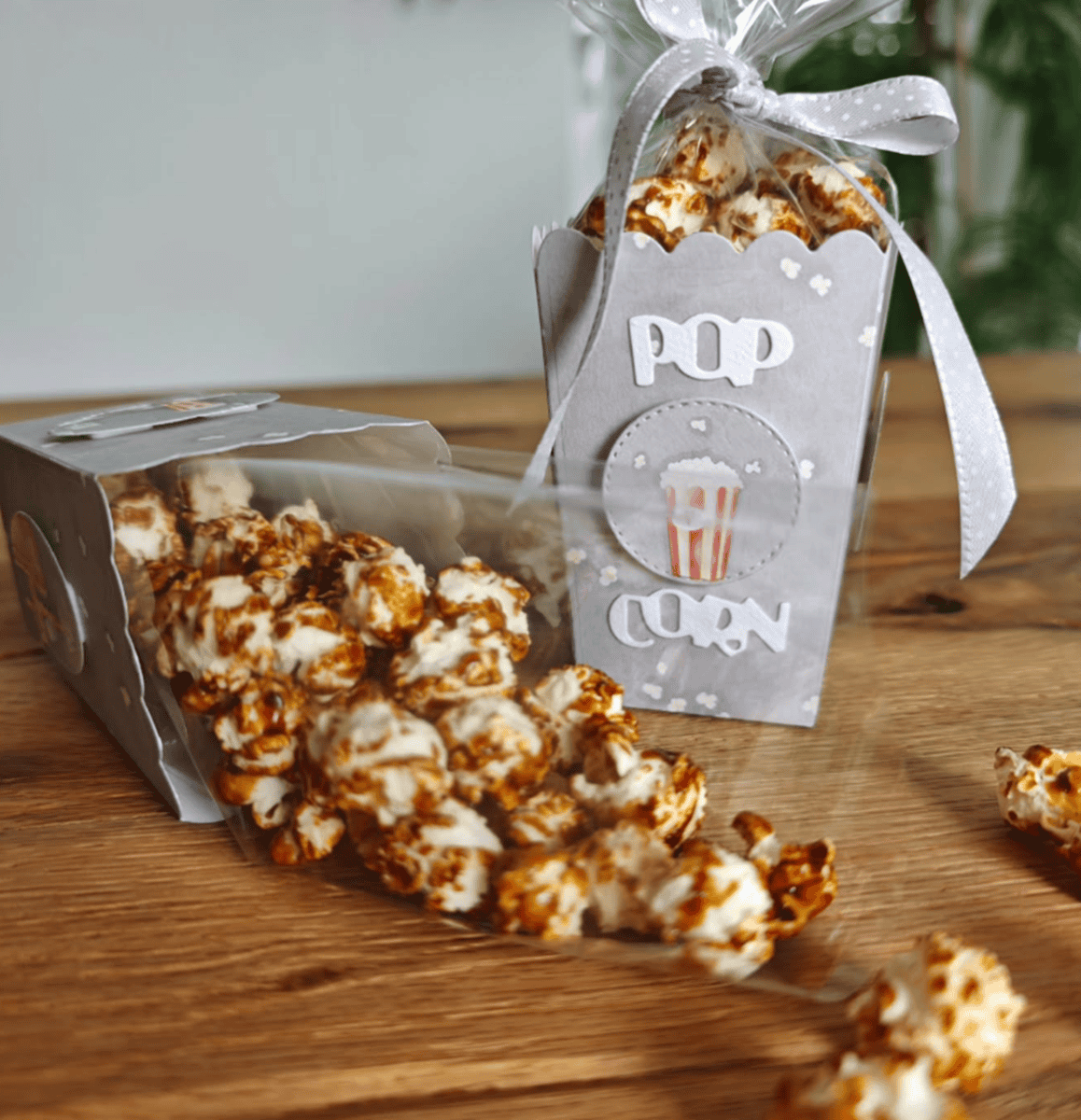 Zwei Stanzschablone Popcornschachteln von Stanzenshop.de - eine aufrecht als Gastgeschenke Hochzeit mit "POP CORN"-Etikett und Schleife, die andere umgekippt mit Karamell-Popcorn, das auf eine Holzfläche spritzt.