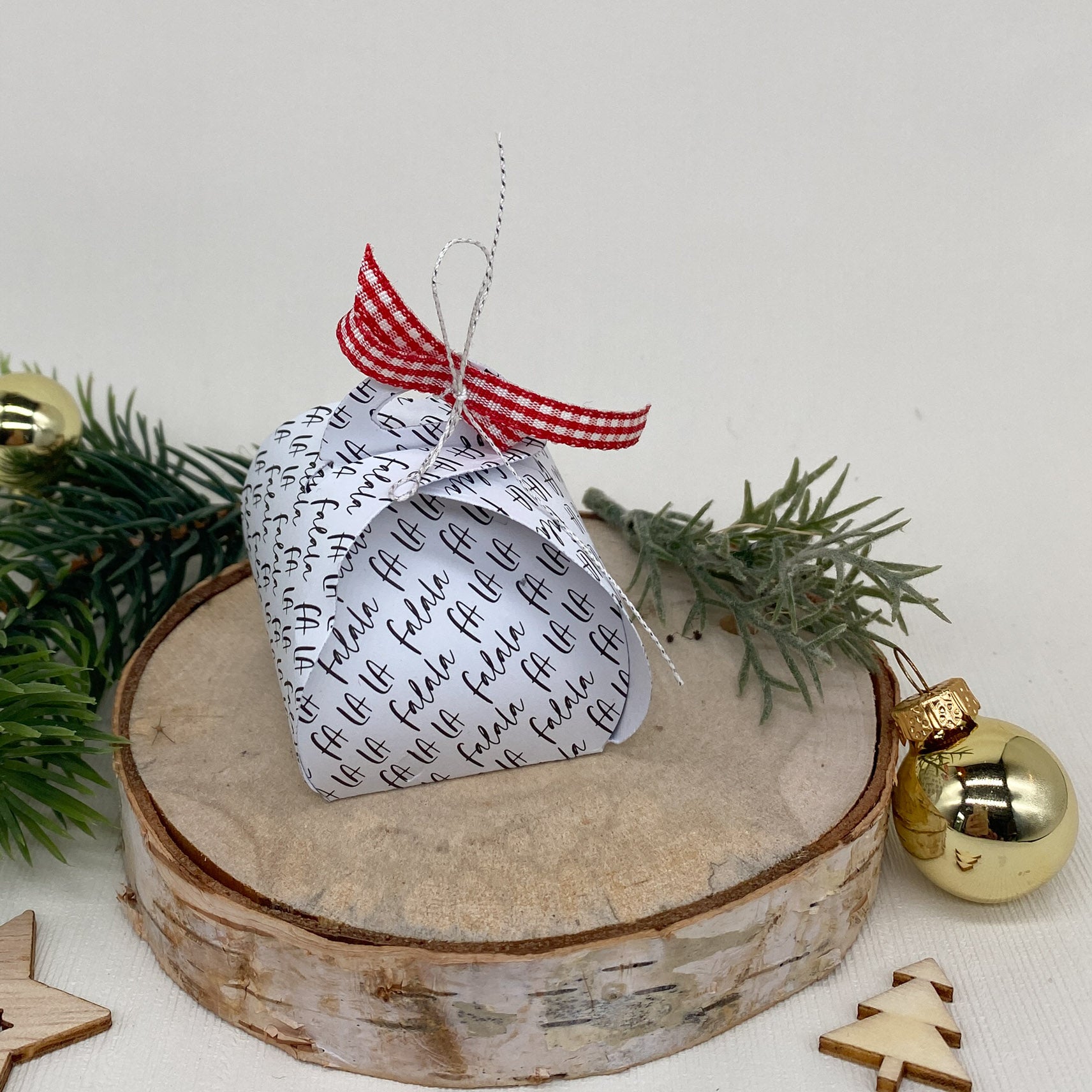 Ein Stanzschablone Schmuckkästchen von Stanzenshop.de mit schwarzem Text und einem rot karierten Band sitzt auf einer Holzplatte, umgeben von Tannenzweigen, einem goldenen Ornament und Holzdekorationen.