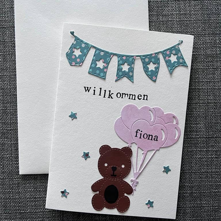 Eine handgefertigte Grußkarte mit einem Bären aus der Stanzschablone Teddybär von Stanzenshop.de, der drei lila Luftballons mit der Aufschrift "Fiona", ein blaues Banner und den Text "Willkommen" in der Hand hält - ideal für Scrapbooking oder Basteln. Inklusive Umschlag.