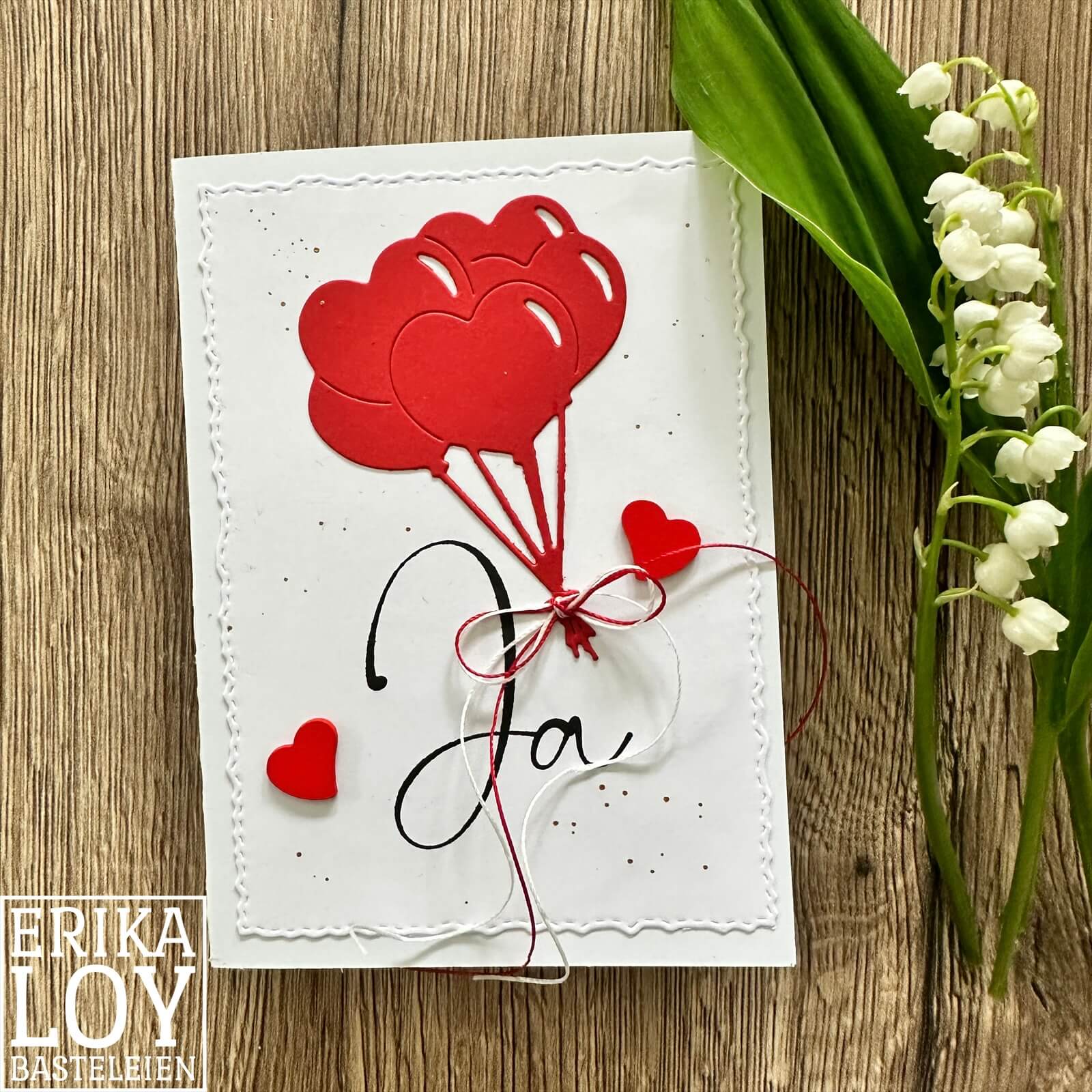 Handgemachte Karte mit zwei roten Herzluftballons aus der "Stanzschablone: Bund Luftballons in Herzform" von Stanzenshop.de, mit "Ja" in Schreibschrift und Maiglöckchen, auf einer Holzunterlage arrangiert für zusätzlichen Charme.