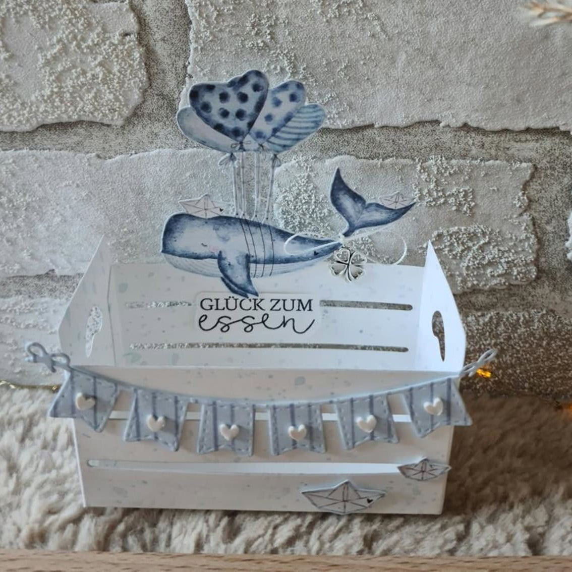 Die Stanzschablone: Großer Karton von Stanzenshop.de ist eine dekorative weiße Geschenkbox Hochzeit mit einem Wal, herzförmigen Luftballons und dem Text "Glück zum essen" auf der Vorderseite, die vor einem strukturierten Steinmauer-Hintergrund steht.