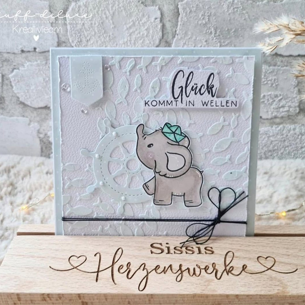 Eine handgefertigte Grußkarte mit Stanzschablone Anker und Steuerrad von Stanzenshop.de, deutschem "Glück kommt in Wellen"-Text, maritimen Stanzelementen und dekorativen Details, präsentiert auf einem Holzständer mit der Aufschrift "Sissis Herzenswerke".