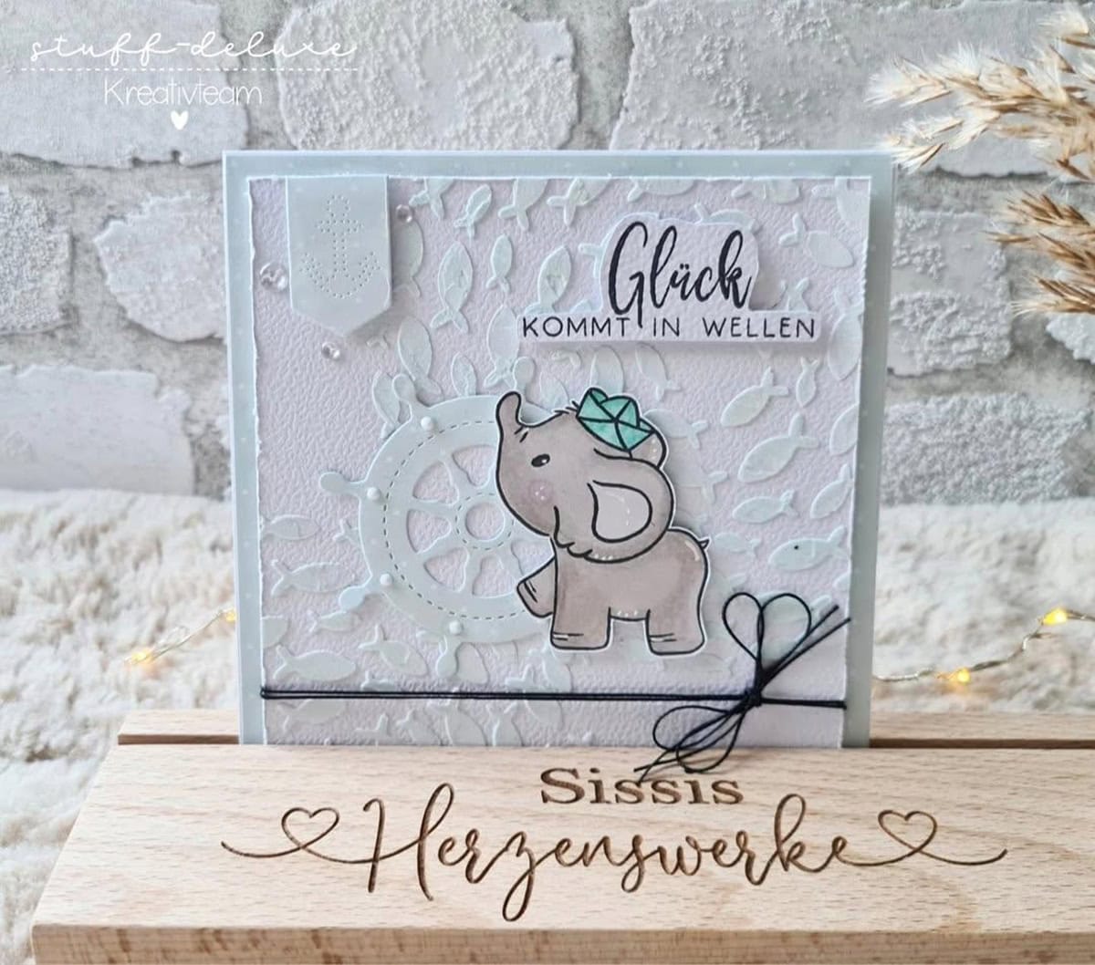 Eine handgefertigte Grußkarte mit Stanzschablone Anker und Steuerrad von Stanzenshop.de, deutschem "Glück kommt in Wellen"-Text, maritimen Stanzelementen und dekorativen Details, präsentiert auf einem Holzständer mit der Aufschrift "Sissis Herzenswerke".