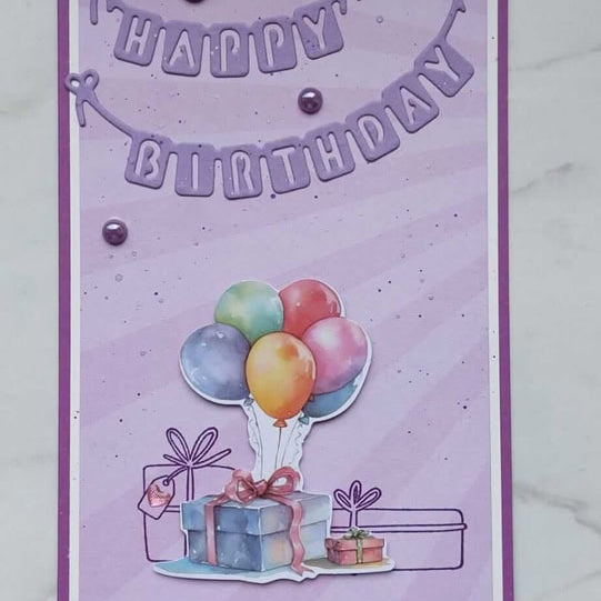Eine handgefertigte Geburtstagskarte mit der Stanzschablone Wimpelkette Happy Birthday von Stanzenshop.de, mit einem festlichen Banner, Luftballons und Geschenkbildern auf einem lila-weißen Hintergrund.