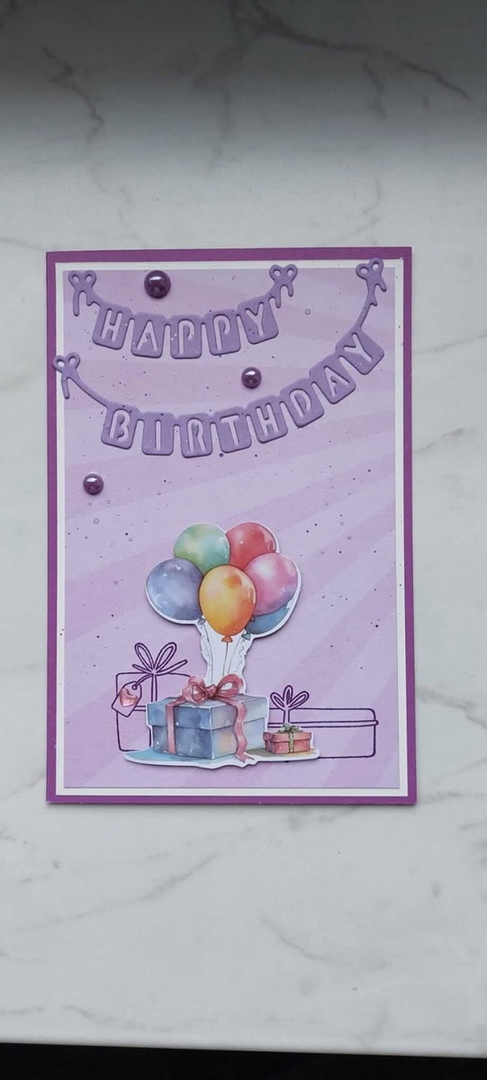 Eine handgefertigte Geburtstagskarte mit der Stanzschablone Wimpelkette Happy Birthday von Stanzenshop.de, mit einem festlichen Banner, Luftballons und Geschenkbildern auf einem lila-weißen Hintergrund.