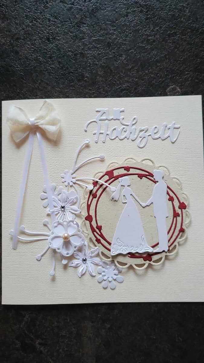 Eine handgefertigte Hochzeitskarte mit einer weißen Schleife, floralen Akzenten und der Silhouette des Brautpaares. Erstellt mit Stanzenshop.de's Stanzschablone Brautpaar, das Wort "Hochzeit" erscheint am oberen Rand.