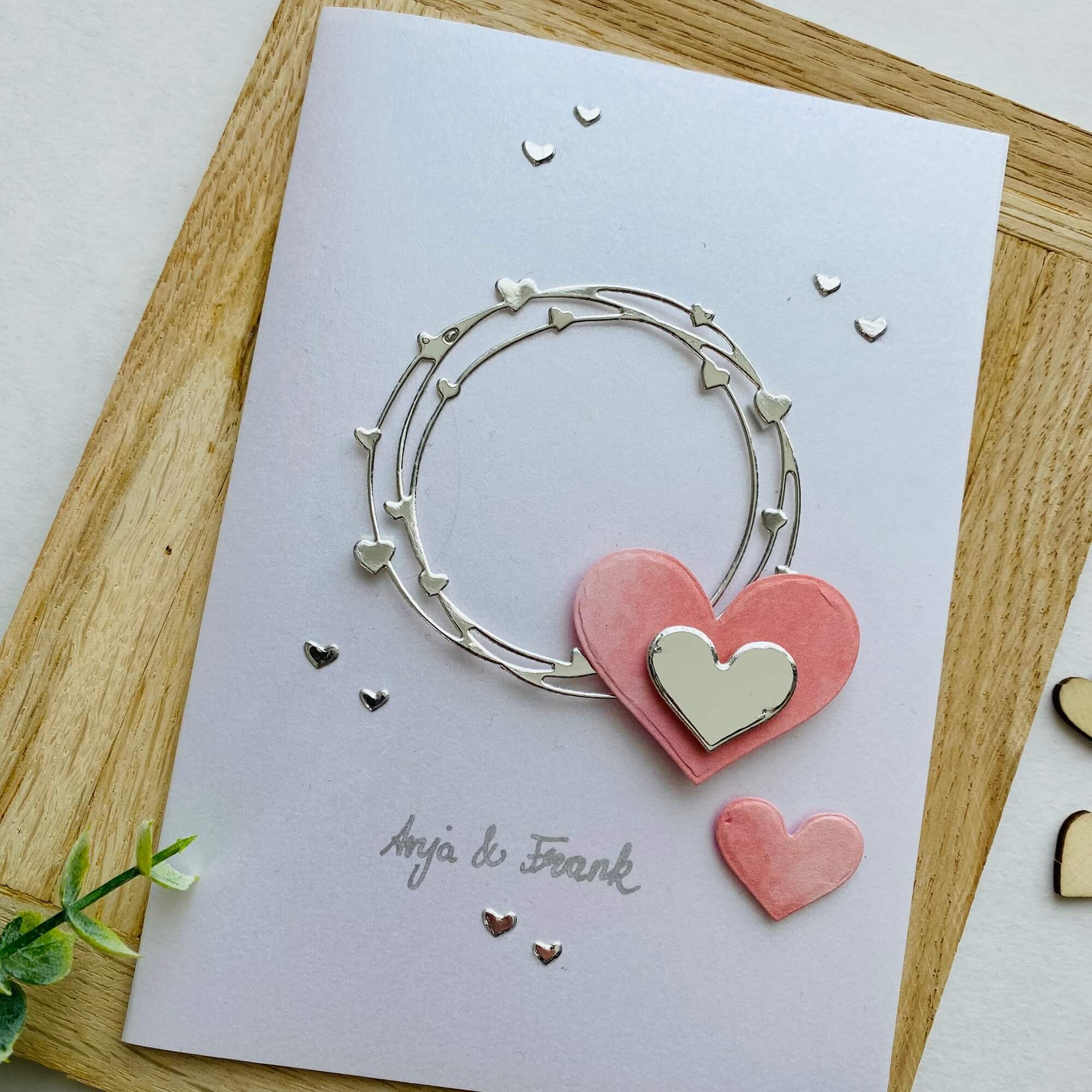 Eine weiße Grußkarte mit einem silbernen und rosa Herzkranz aus der Stanzschablone Herzkranz von Stanzenshop.de mit "Anja & Frank" in Schreibschrift am unteren Rand, perfekt zum Basteln oder Scrapbooking.