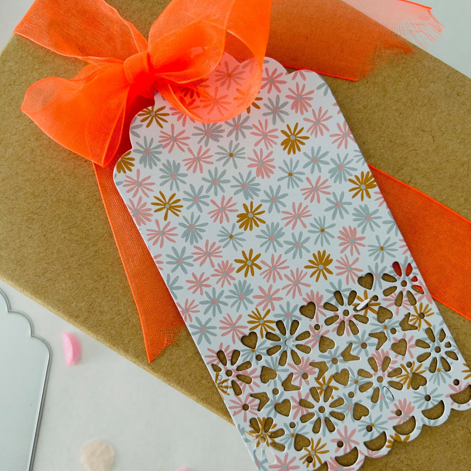 Die Stanzschablone Anhänger mit Verzierung von Stanzenshop.de ist eine braune Geschenkbox mit orangefarbener Schleife und einem floralen gold-pinken Anhänger, ideal für Scrapbooking oder zur Verwendung mit einem Big Shot.