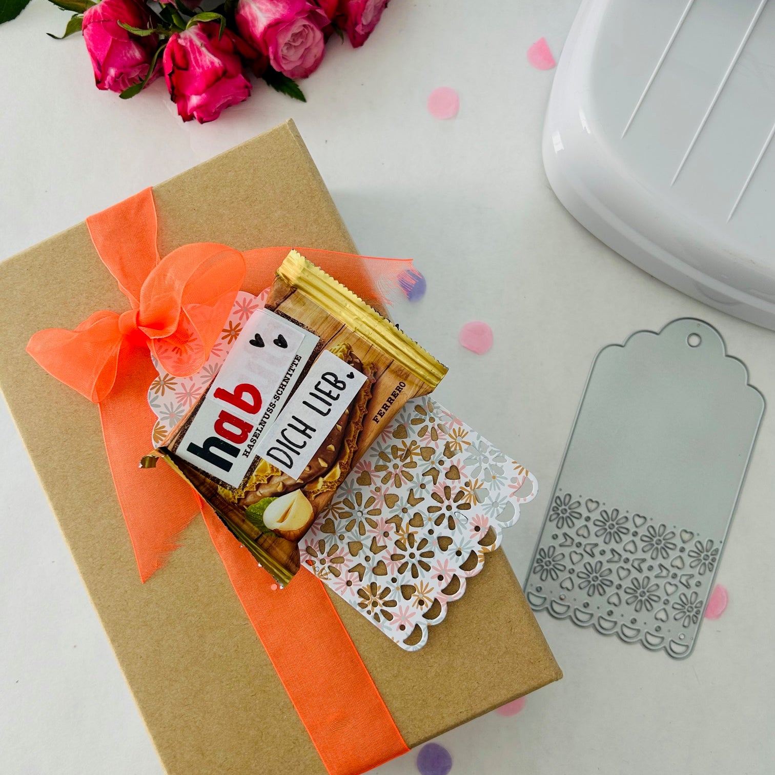 Eine Geschenkschachtel mit orangefarbenem Band, Pralinen, Danksagung, rosa Rosen, einer Stanzschablone Anhänger mit Verzierung von Stanzenshop.de (ideal für Scrapbooking) und einem dekorativen Anhänger steht auf einer weißen Fläche.