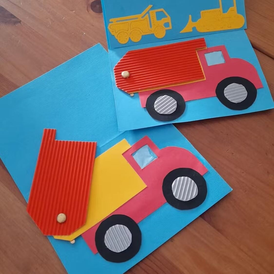 Zwei handgefertigte Karten mit Stanzschablone Baufahrzeuge von Stanzenshop.de, mit Kippermotiven aus farbigem Papier und Wellpappe. Eine Karte ist offen und zeigt zusätzliche Fahrzeugformen auf einem Holztisch - ideal für Basteln-Fans.