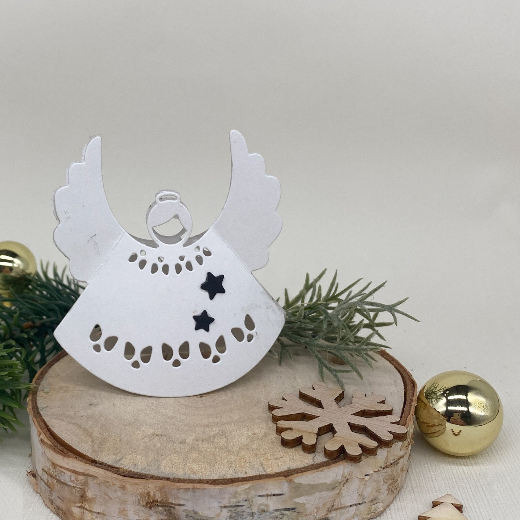 Die Stanzschablone: Engel von Stanzenshop.de ist eine weiße dekorative Engelstanze, ideal für Scrapbooking oder Bastelprojekte, abgebildet auf Holz mit einer Schneeflocke, Baum, Tannenzweig und Goldkugeln.