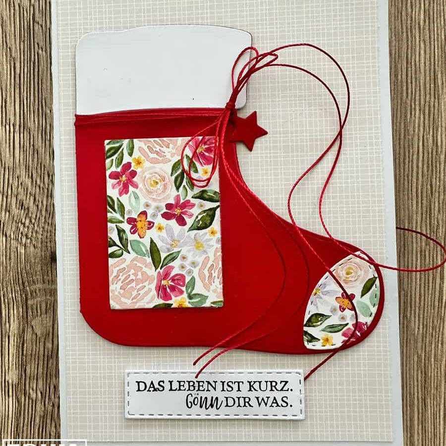 Handgefertigte Grußkarte mit einem roten Weihnachtsstrumpf, hergestellt mit Stanzschablone: Stiefelumschlag von Stanzenshop.de und floralen Motiven. Perfekt für Bastelprojekte Weihnachten. Inklusive roter Schnur und Anhänger: "DAS LEBEN IST KURZ. GÖNN DIR WAS.