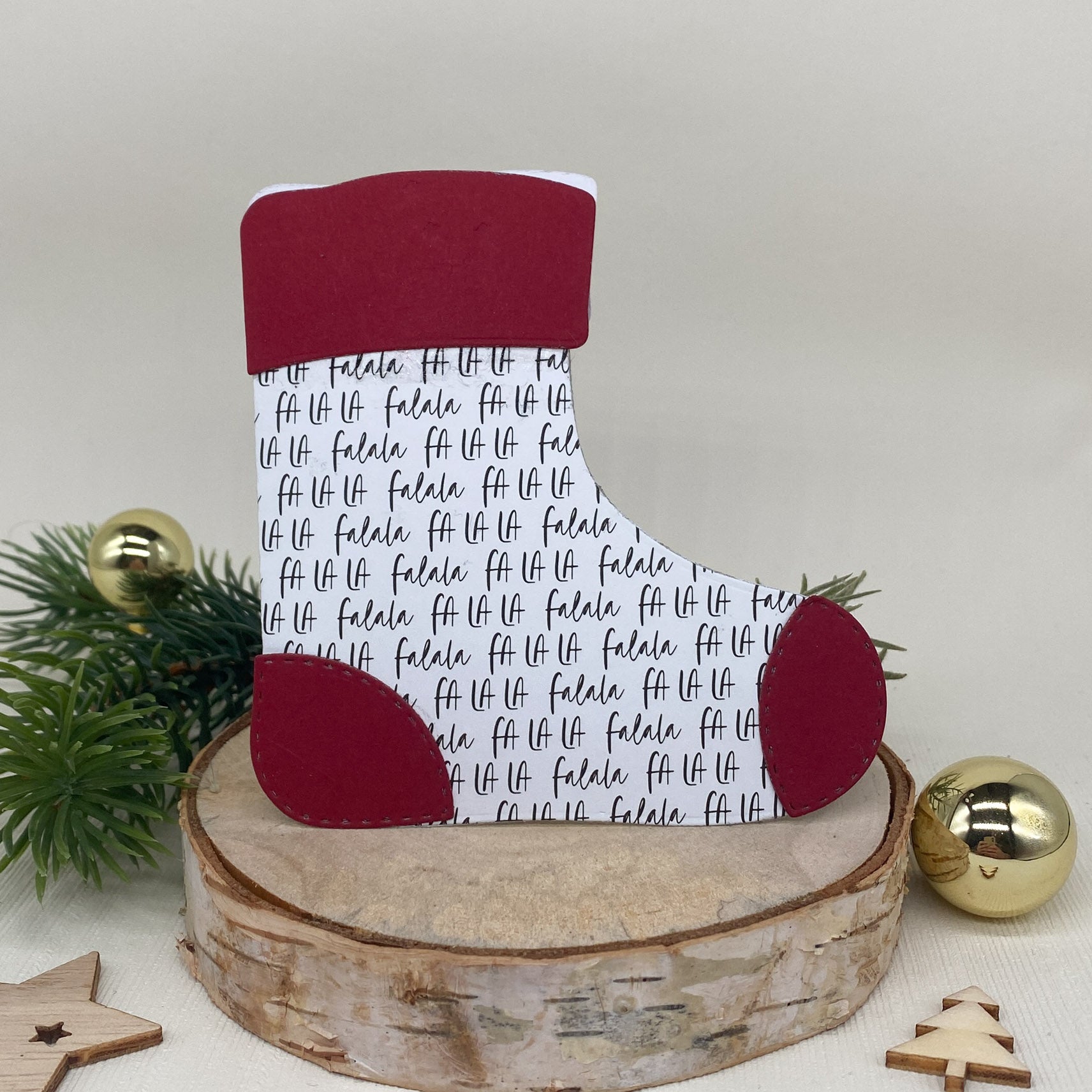 Ein festlicher Papierstrumpf, der mit der Stanzschablone gefertigt wurde: Stiefelumschlag von Stanzenshop.de und dem Text "fa la la", mit roten Akzenten, ruht auf einer Holzscheibe inmitten von Tannenzweigen und goldenen Ornamenten - ideal für Weihnachts-Bastelprojekte.