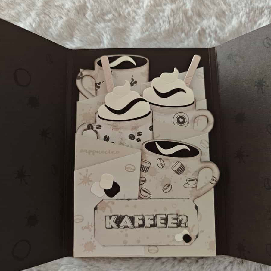 Eine handgemachte Karte mit geschichteten Papierausschnitten von Kaffeetassen unter Verwendung der "Stanzschablone: Tassen" von Stanzenshop.de, gekrönt mit Schlagsahne und "KAFFEE" auf einem strukturierten Hintergrund.