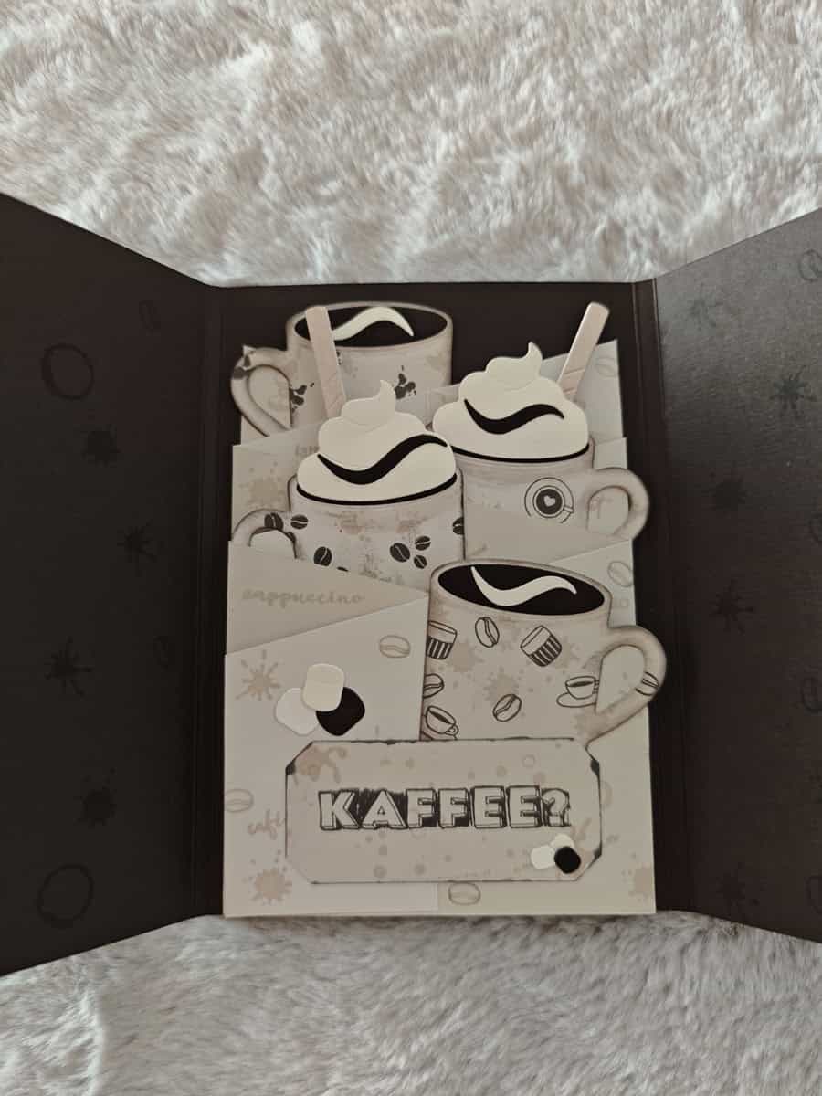 Eine handgemachte Karte mit geschichteten Papierausschnitten von Kaffeetassen unter Verwendung der "Stanzschablone: Tassen" von Stanzenshop.de, gekrönt mit Schlagsahne und "KAFFEE" auf einem strukturierten Hintergrund.