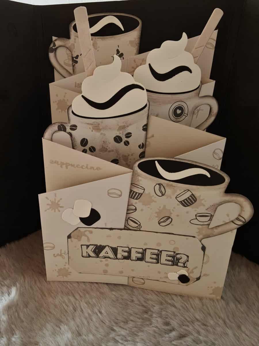 Eine handgefertigte Karte mit geschichteten Kaffeetassenausschnitten und "Kaffee" auf der Vorderseite, hergestellt mit der Stanzschablone: Tassen von Stanzenshop.de als kreatives Bastelzubehör, dargestellt auf einer pelzigen Oberfläche.
