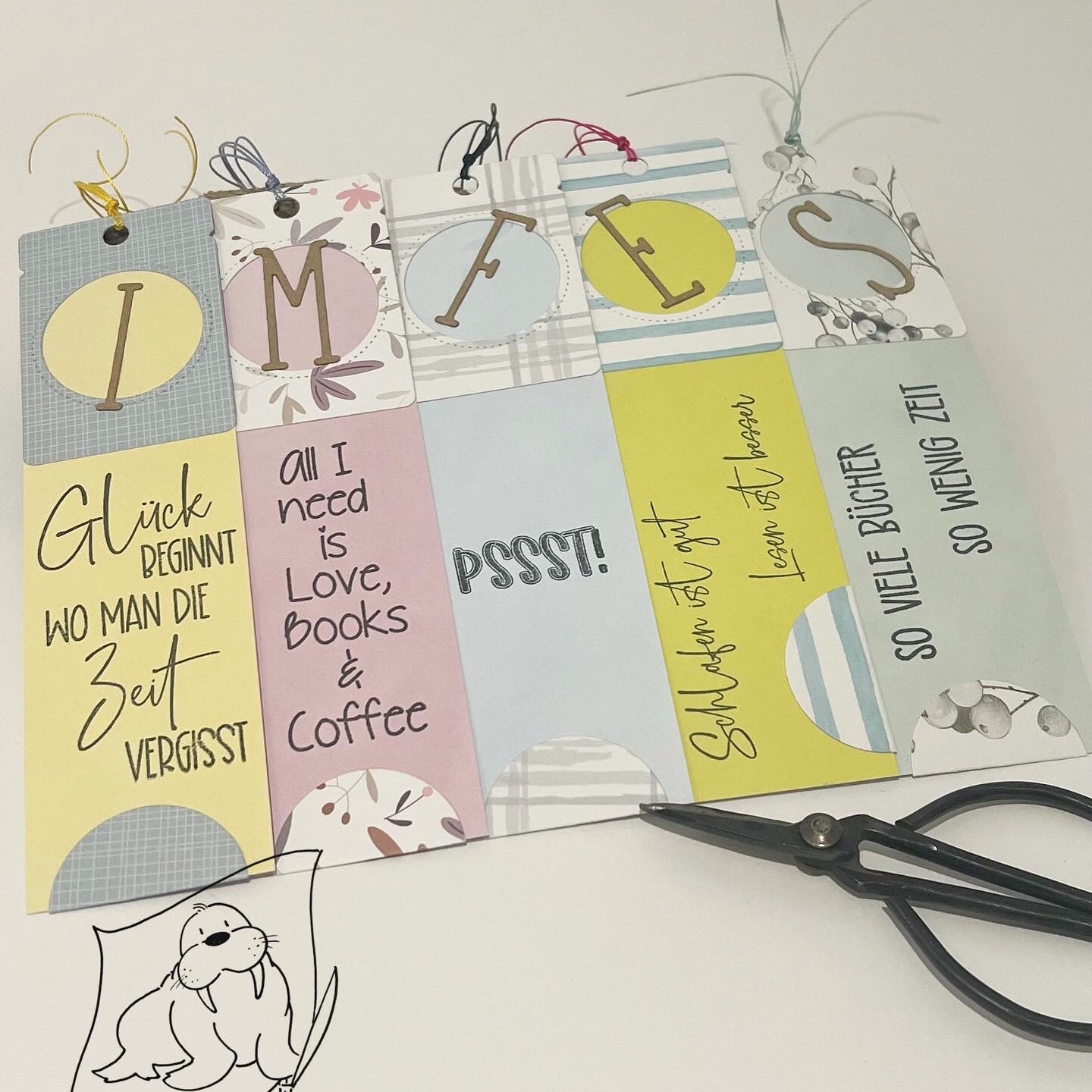 Die Stanzschablone: Klappetikett von Stanzenshop.de enthält sechs verzierte Lesezeichen mit deutschen und englischen Zitaten, ideal für Scrapbooking, abgebildet neben einer schwarzen Schere und einer Zeichnung einer Robbe mit Schere.