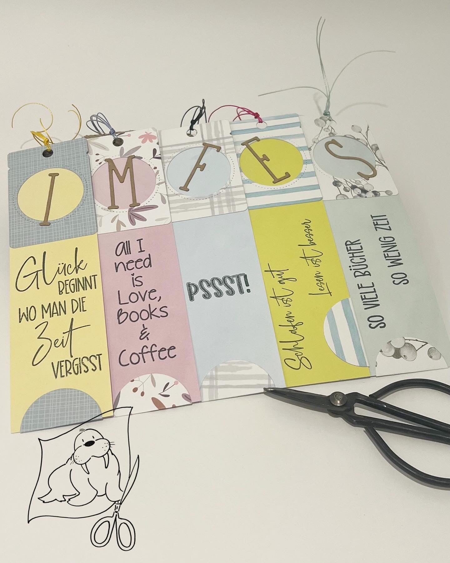 Die Stanzschablone: Klappetikett von Stanzenshop.de enthält sechs verzierte Lesezeichen mit deutschen und englischen Zitaten, ideal für Scrapbooking, abgebildet neben einer schwarzen Schere und einer Zeichnung einer Robbe mit Schere.