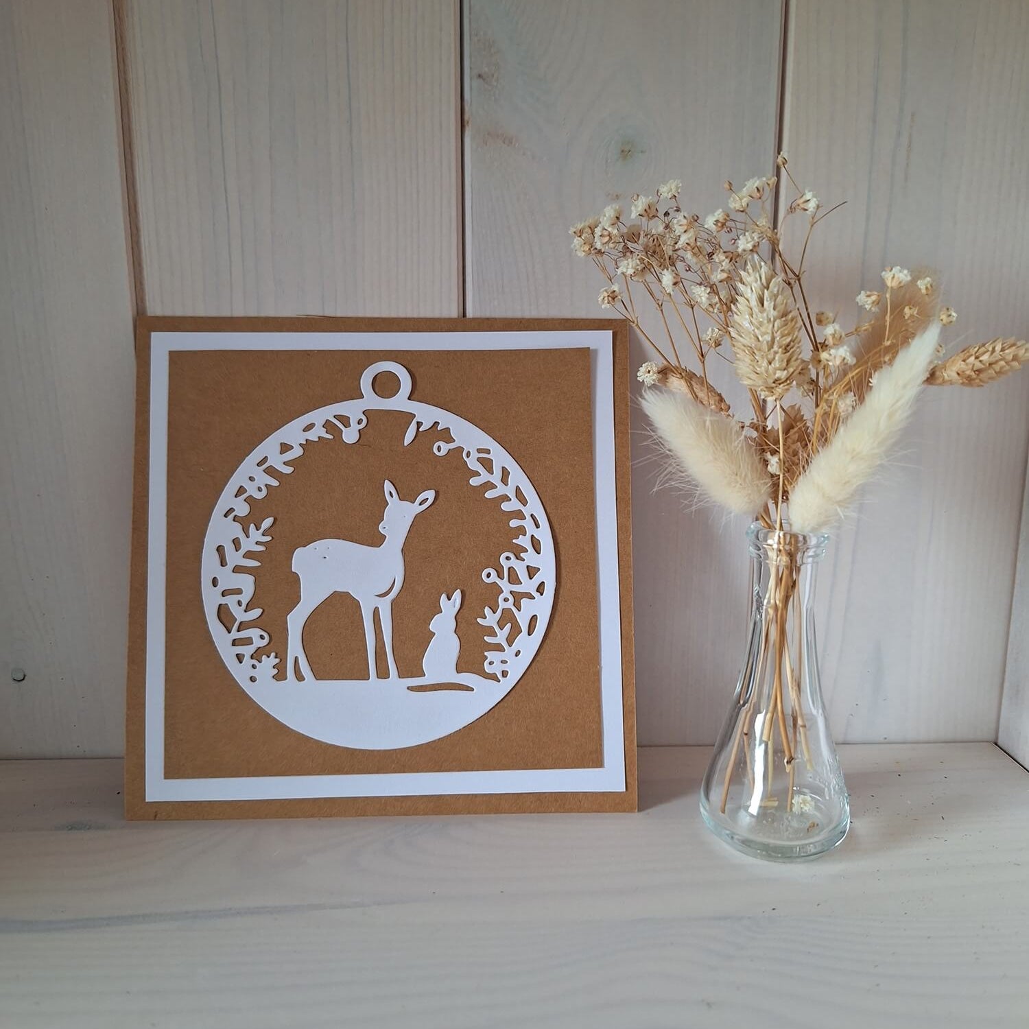 Eine braun-weiße Grußkarte aus der Stanzschablone: Reh mit Hase von Stanzenshop.de sitzt neben einer Vase mit getrockneten Blumen auf einer hellen Holzoberfläche - ideal für Bastelprojekte oder Scrapbooking.