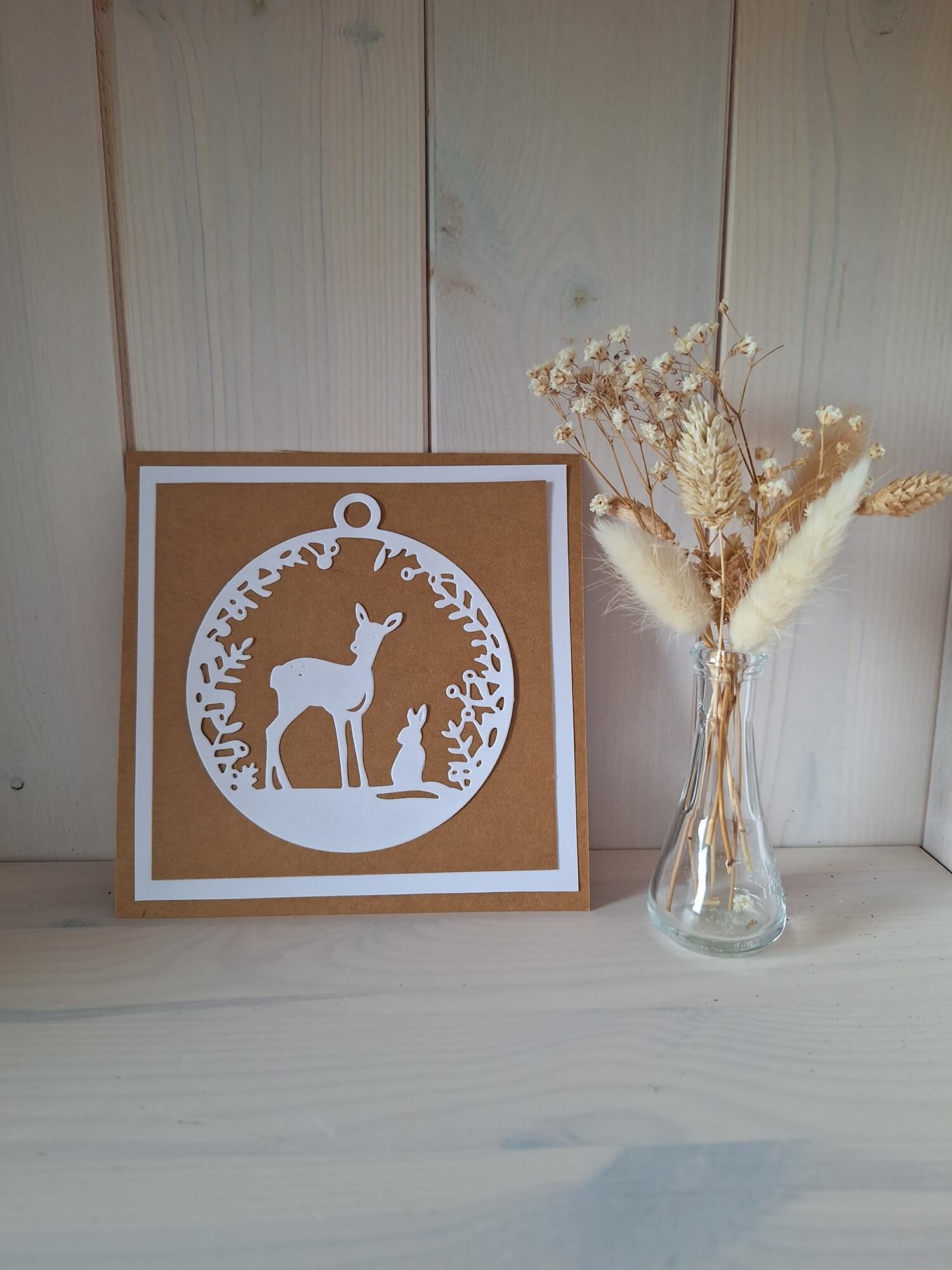 Eine braun-weiße Grußkarte aus der Stanzschablone: Reh mit Hase von Stanzenshop.de sitzt neben einer Vase mit getrockneten Blumen auf einer hellen Holzoberfläche - ideal für Bastelprojekte oder Scrapbooking.
