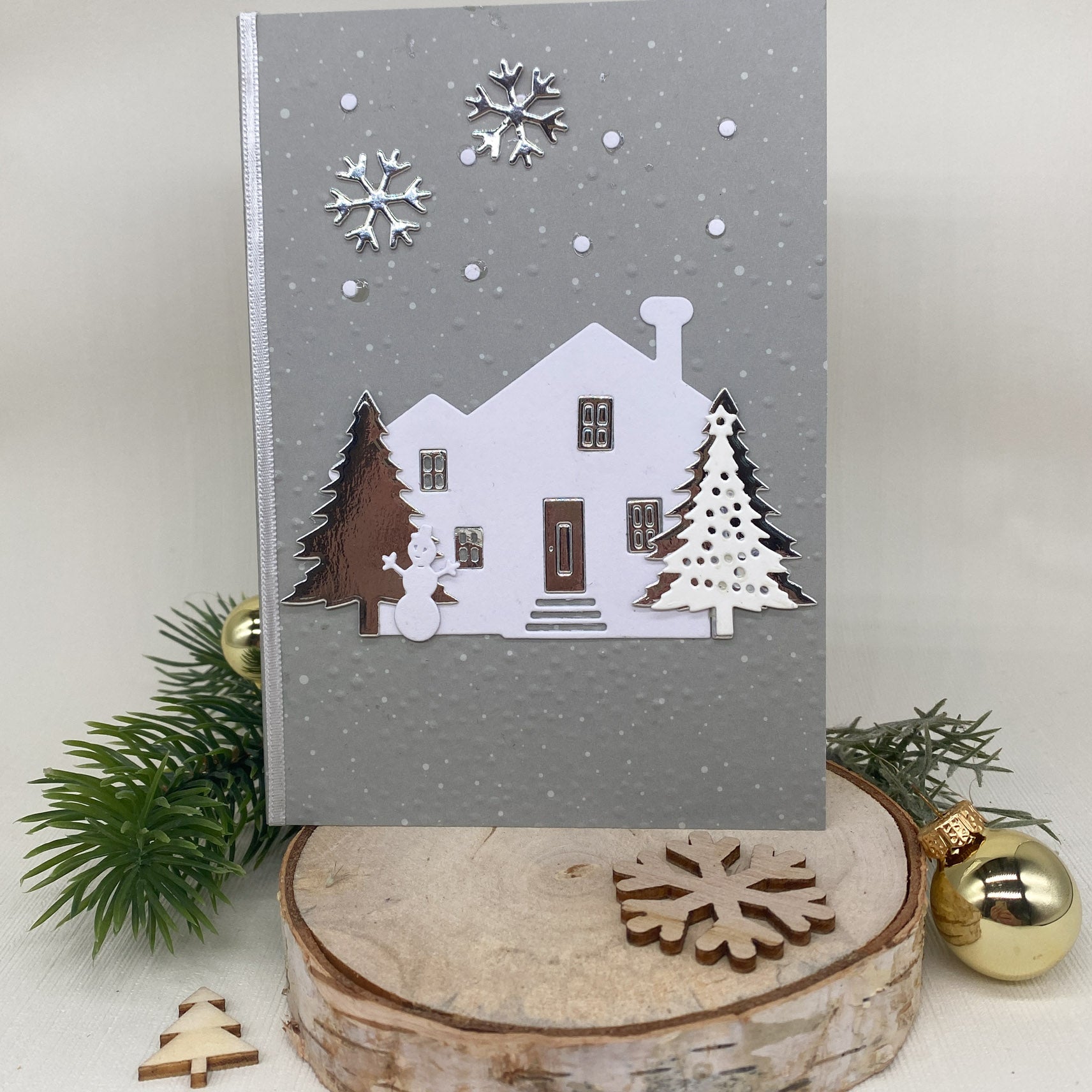 Eine graue Weihnachtskarte mit Stanzschablone: Haus mit Tannen von Stanzenshop.de, mit einem weißen Haus, Bäumen und einem Schneemann inmitten von Schneeflocken auf einem Holzständer mit Tannenzweigen - ideal für Scrapbooking oder Bastelprojekte.