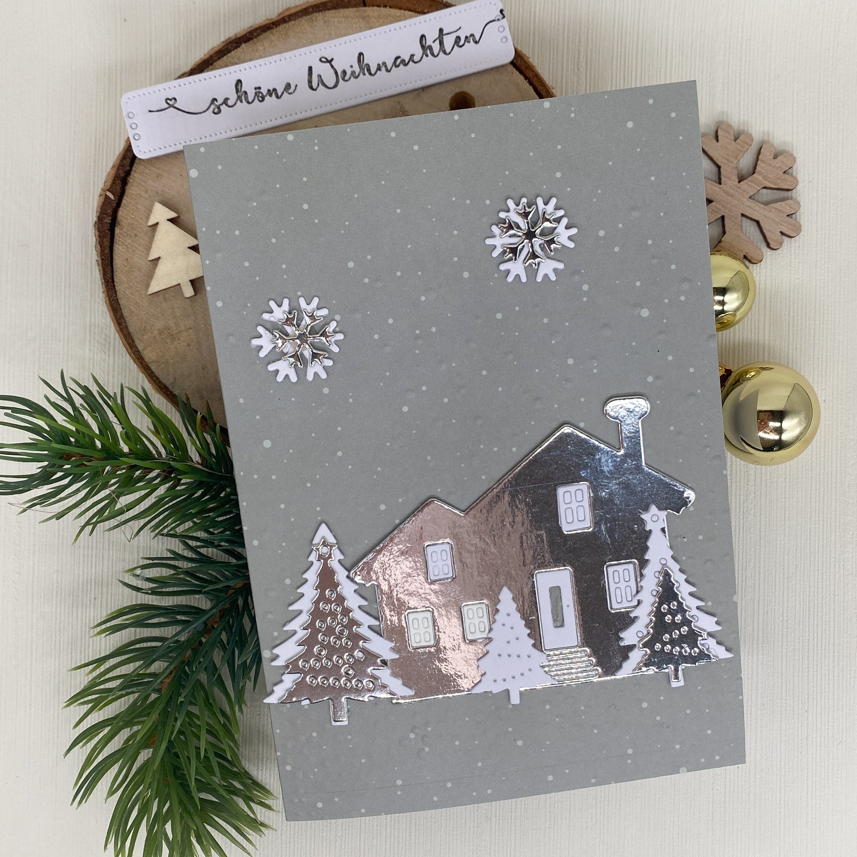 Eine graue Weihnachtskarte mit der Stanzenshop.de Stanzschablone: Haus mit Tannen, mit weißen Bäumen und Schneeflocken, auf einer Holzplatte mit Tannenzweigen, Ornamenten und einem "Schöne Weihnachten" Tag - ideal für Scrapbooking oder Bastelprojekte.