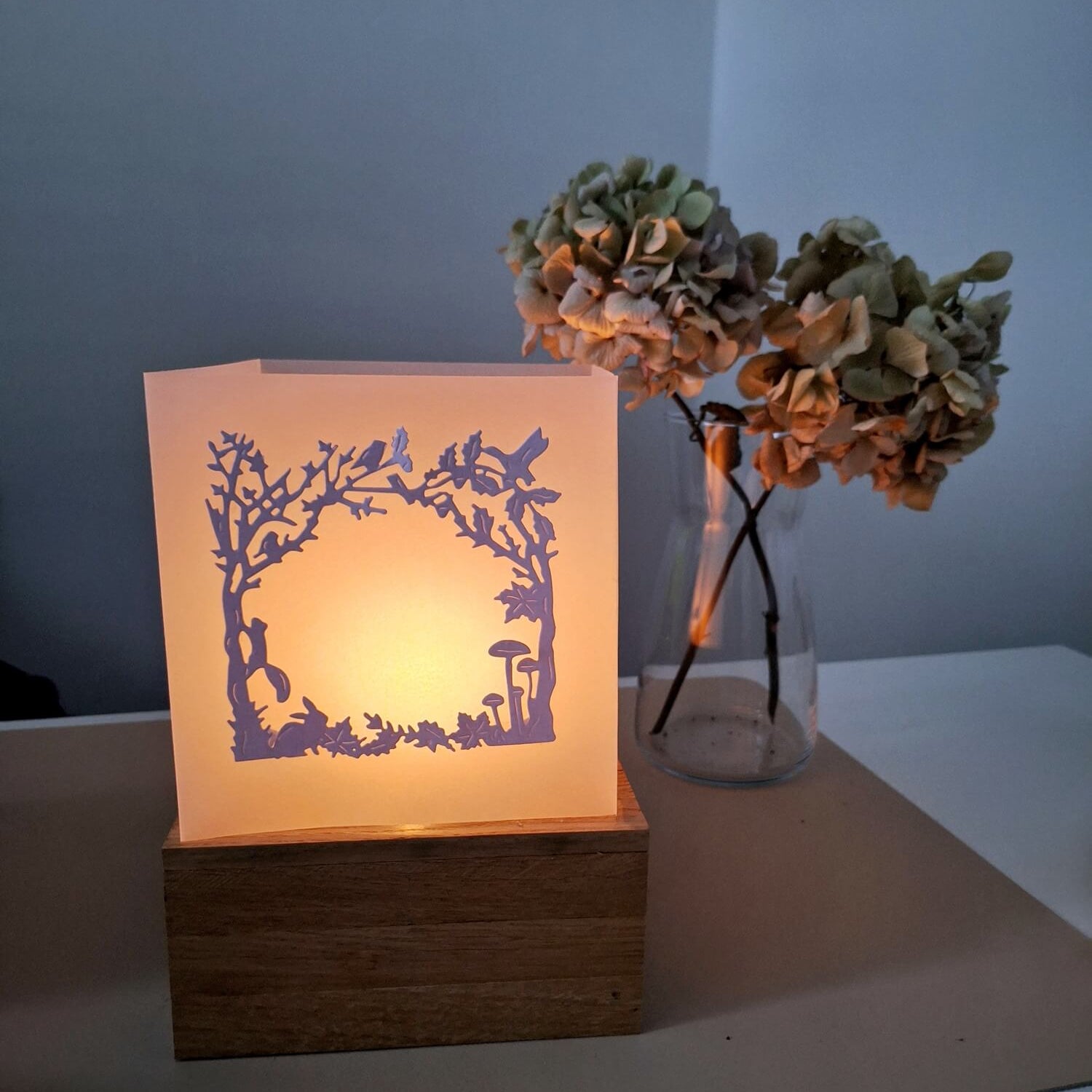 Eine Papierlaterne mit Herbstmotiven leuchtet auf einem Holzsockel neben getrockneten Hortensien und bietet die perfekte Inspiration für Bastelprojekte mit der Stanzschablone: Herbstwald von Stanzenshop.de auf einem weißen Tisch.