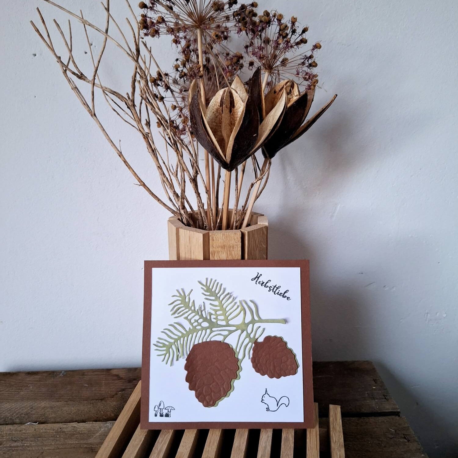 Die "Stanzschablone: Zapfenzweig" von Stanzenshop.de mit Tannenzapfen und -nadeln wird vor einer Vase mit getrockneten Pflanzen auf einer Holzlattenunterlage gezeigt - ideal für Scrapbooking oder Bastelprojekte.