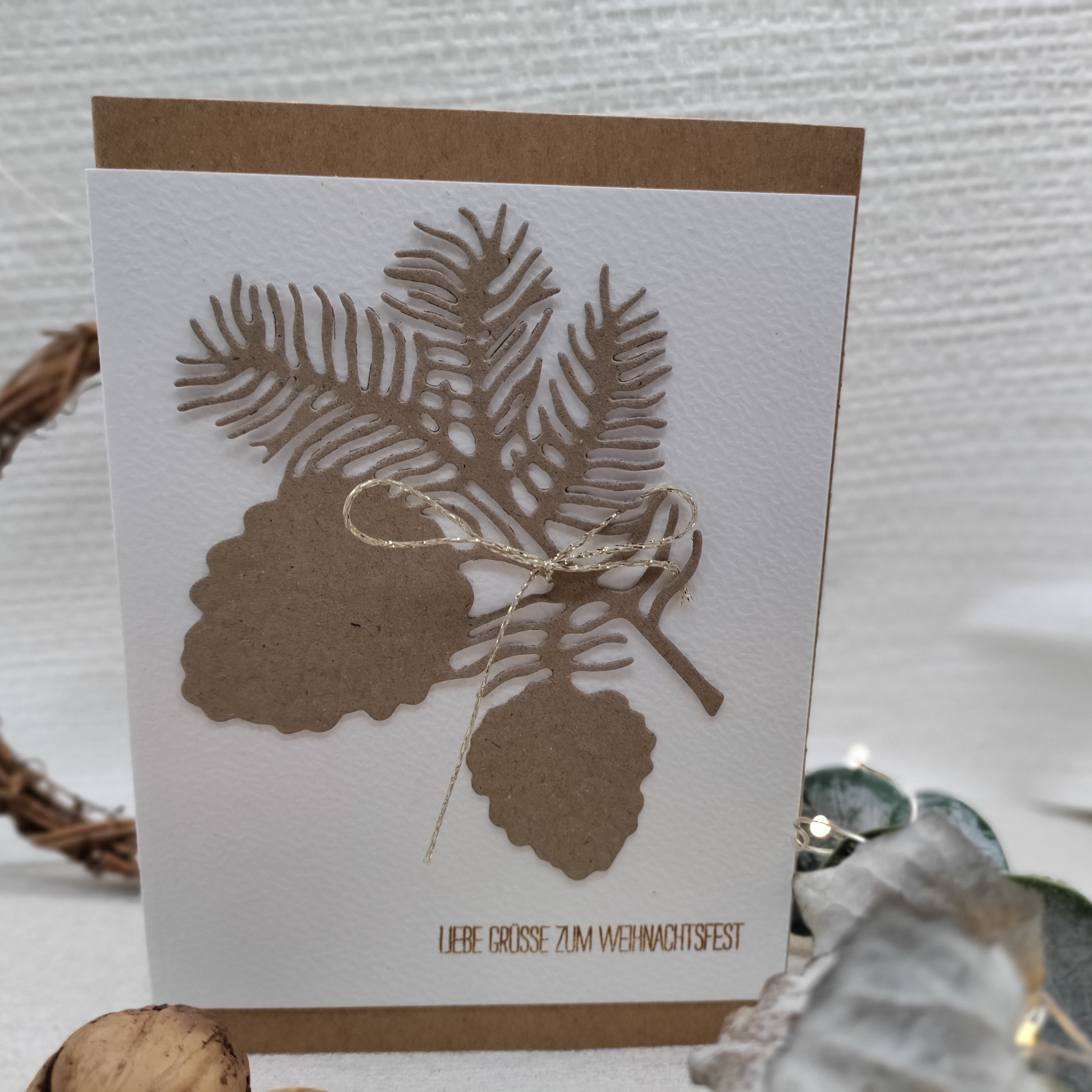 Diese handgefertigte Karte zeigt ausgeschnittene Tannenzweige und Zapfen mit der Stanzschablone: Zapfenzweig von Stanzenshop.de, verziert mit einer goldenen Schleife und dem Schriftzug "Liebe Grüße zum Weihnachtsfest" - ideal zum Basteln oder Scrapbooking.